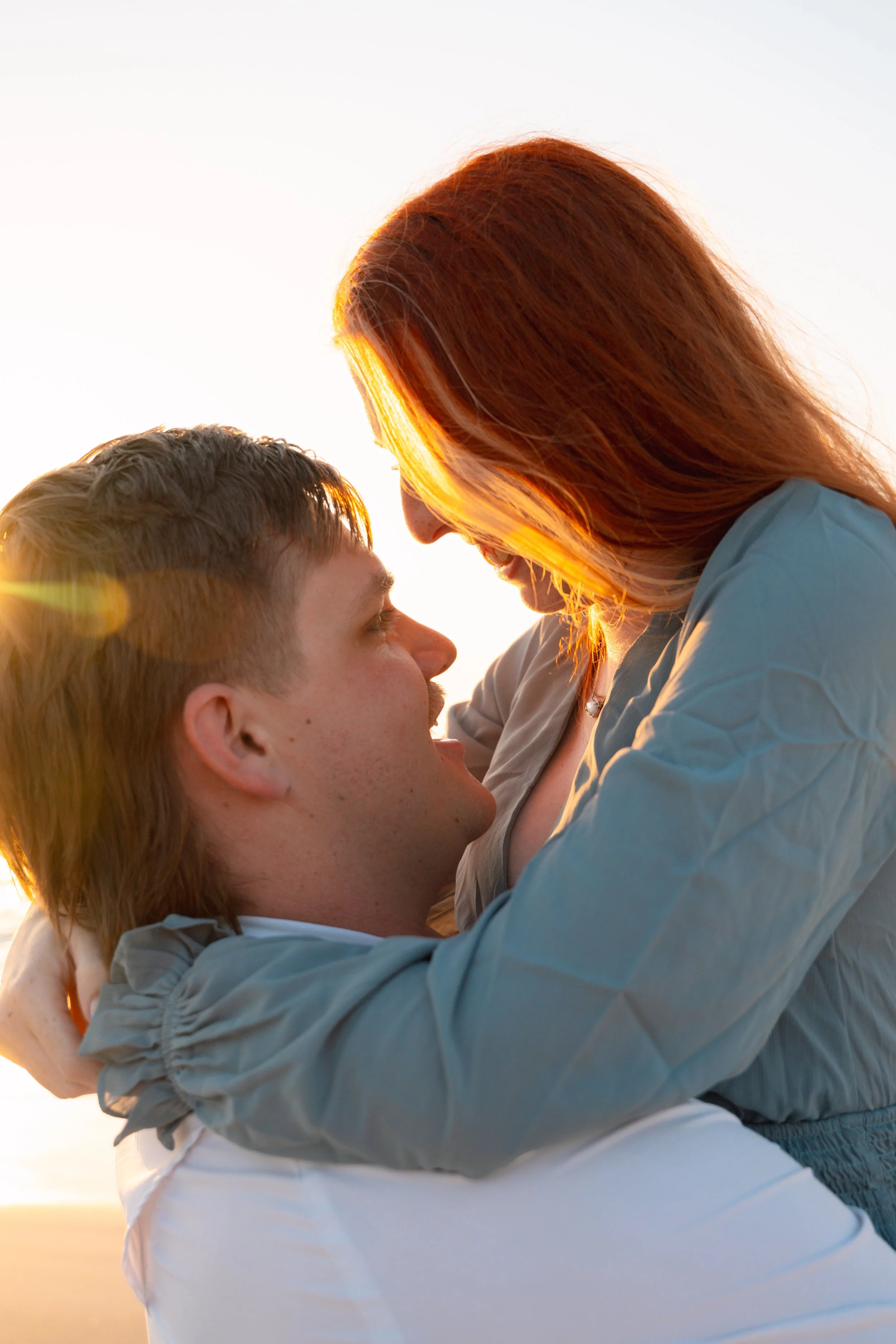A smiling woman with red hair hugging and looking at a man with light brown hair, both close together, during sunset.
