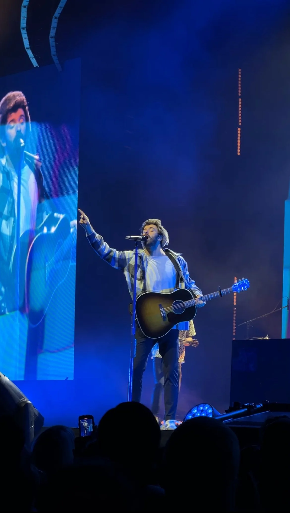 A male musician with a guitar performing on stage, pointing upward with his right hand, in front of a big vertical screen showing his close-up, with blue stage lights and audience silhouettes in the foreground.
