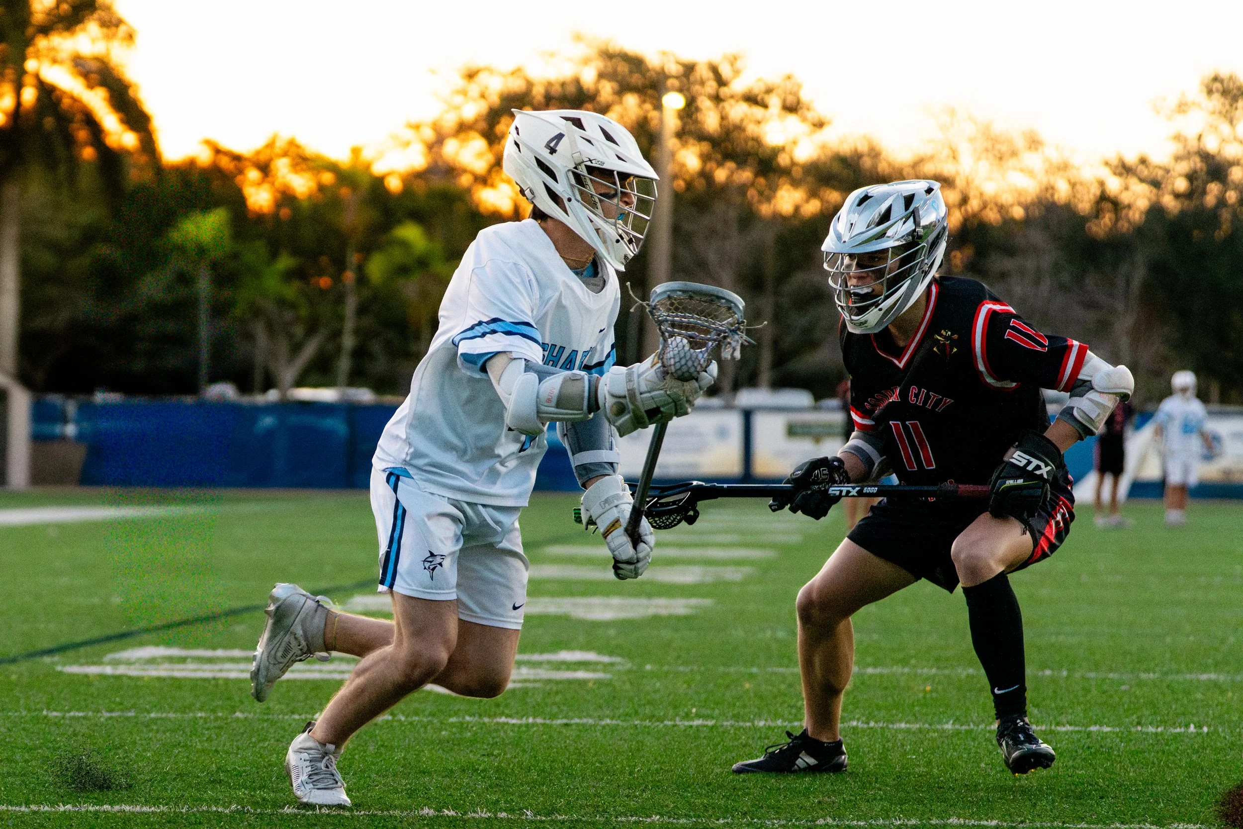 Two lacrosse players wearing helmets and athletic gear compete for possession of the ball on a grassy field during sunset.