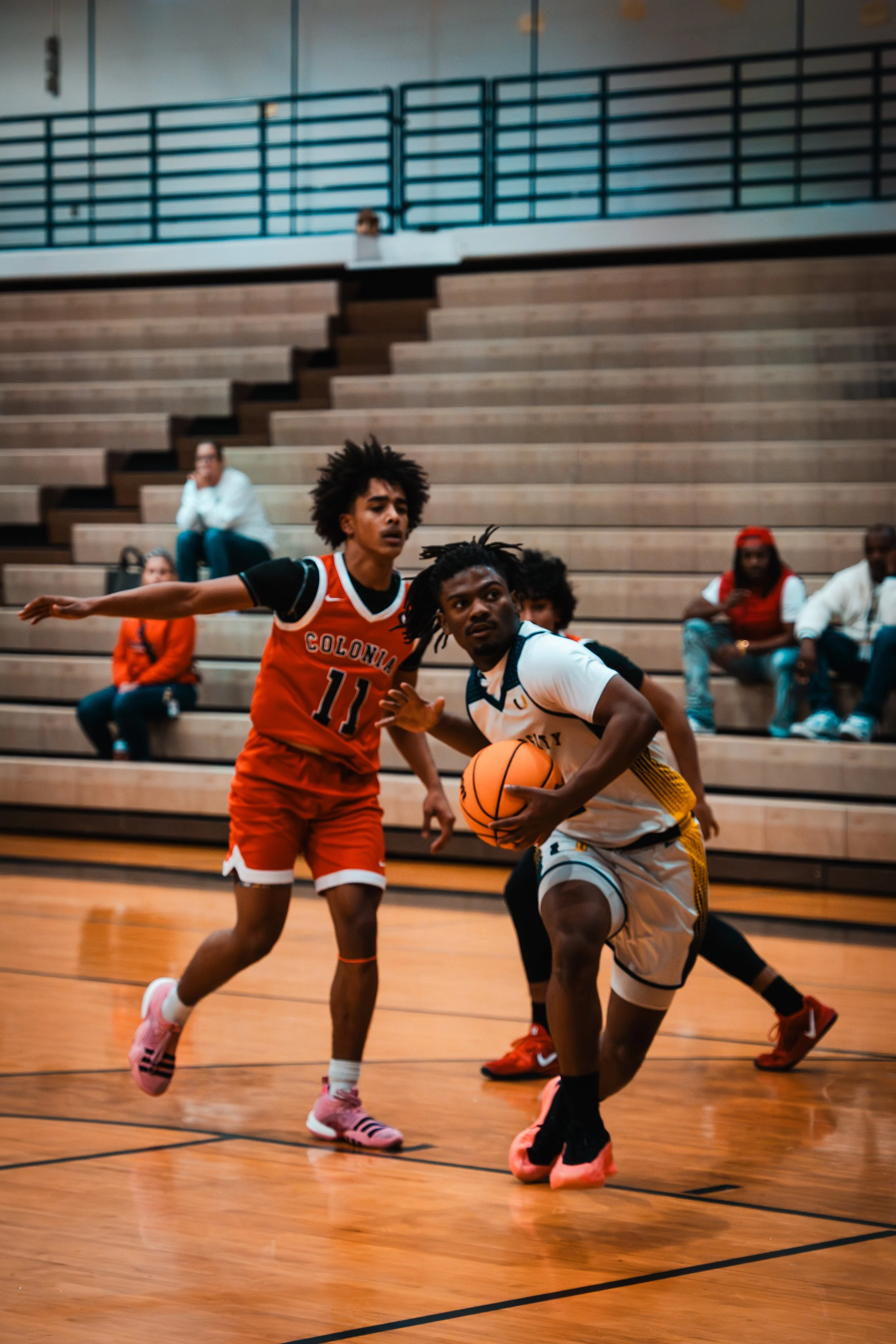 A basketball player in a white and yellow jersey dribbling the ball while a player in a red and black jersey defends him during a game indoors, with spectators sitting on bleachers in the background.