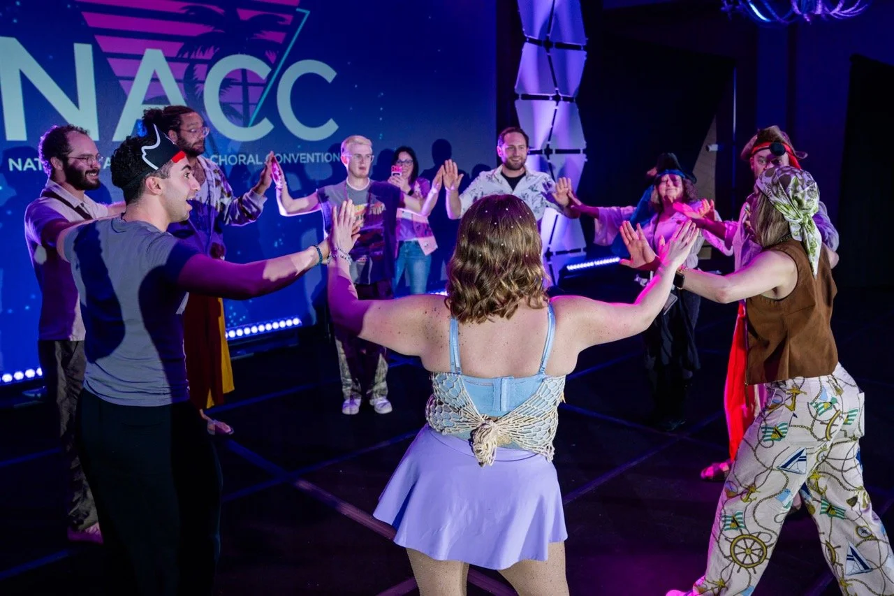 Group of people dancing in a circle on stage during a gathering or event, with a large screen in the background displaying 'NAC'.