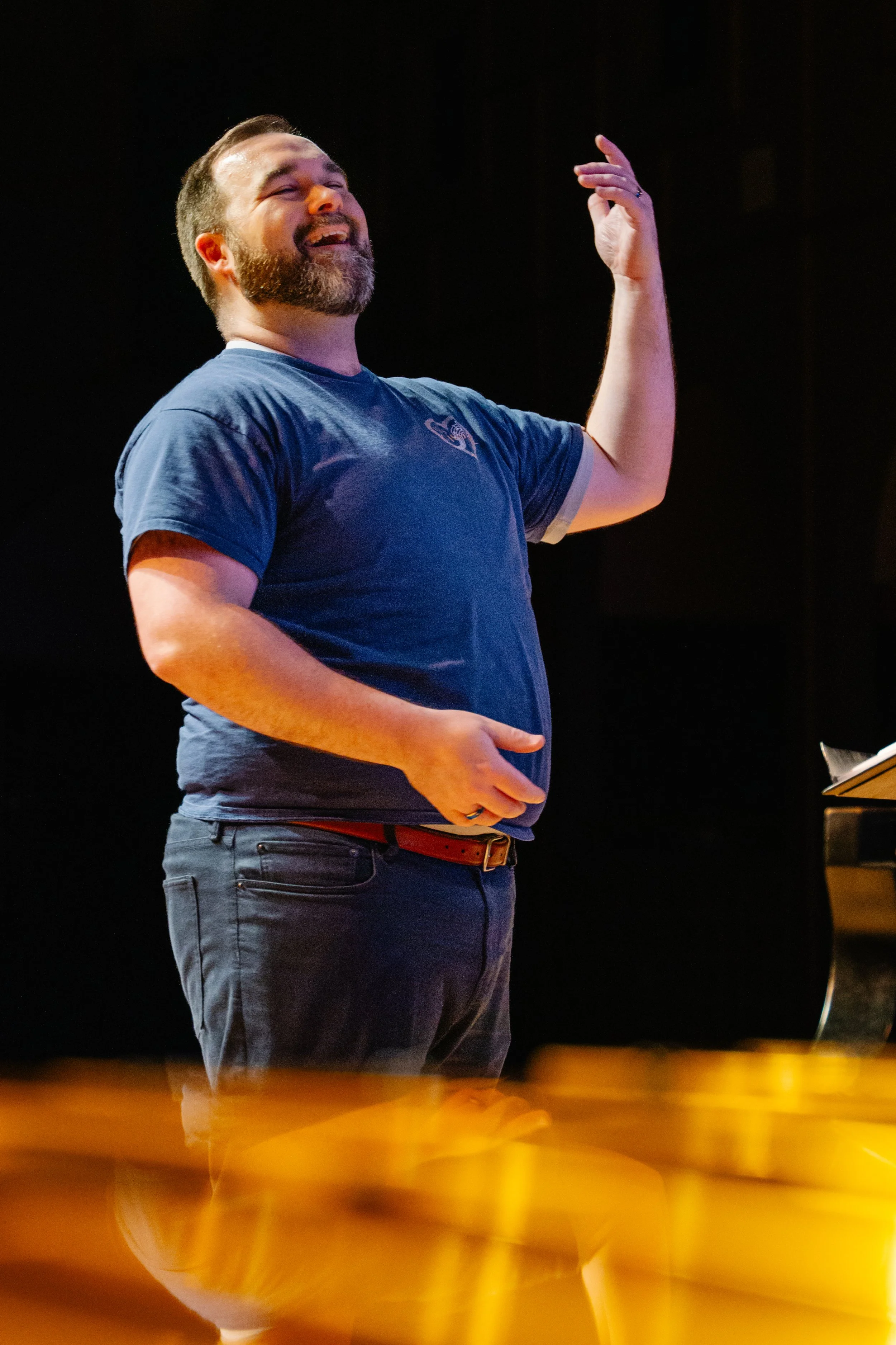 A man with a beard and short hair, wearing a blue t-shirt and dark pants, smiling and gesturing with his right hand.