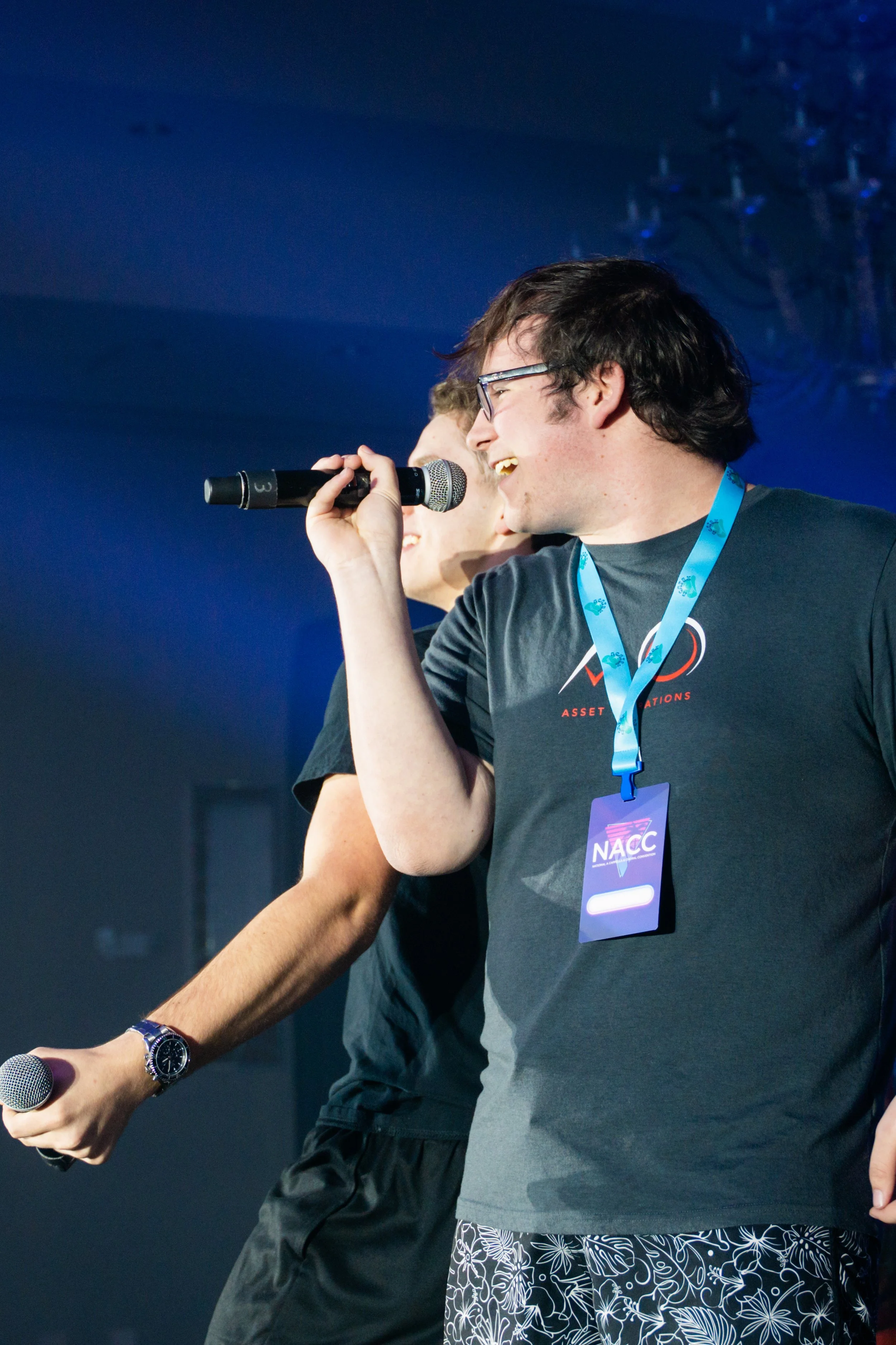 Two young men are smiling and speaking into microphones on stage, with one wearing glasses and a dark t-shirt with a conference badge around his neck, and the other partially visible. The background is dark with stage lighting.