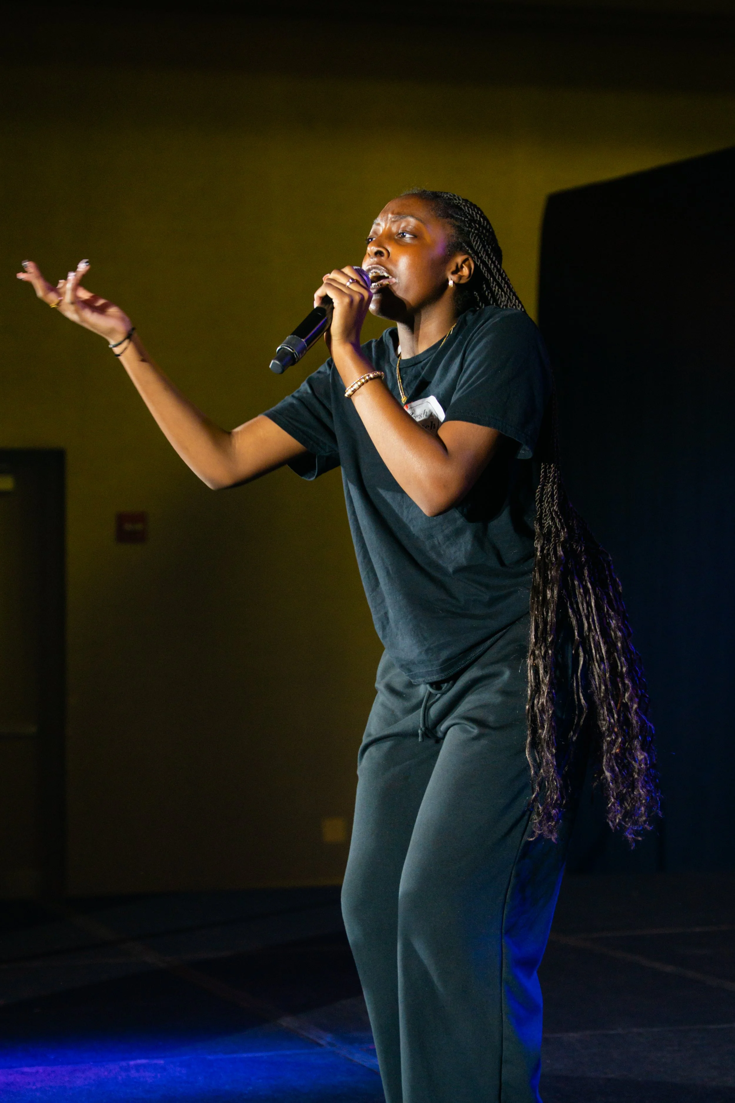 A woman with braided hair wearing a black T-shirt and gray pants singing into a microphone on a stage with dark background.