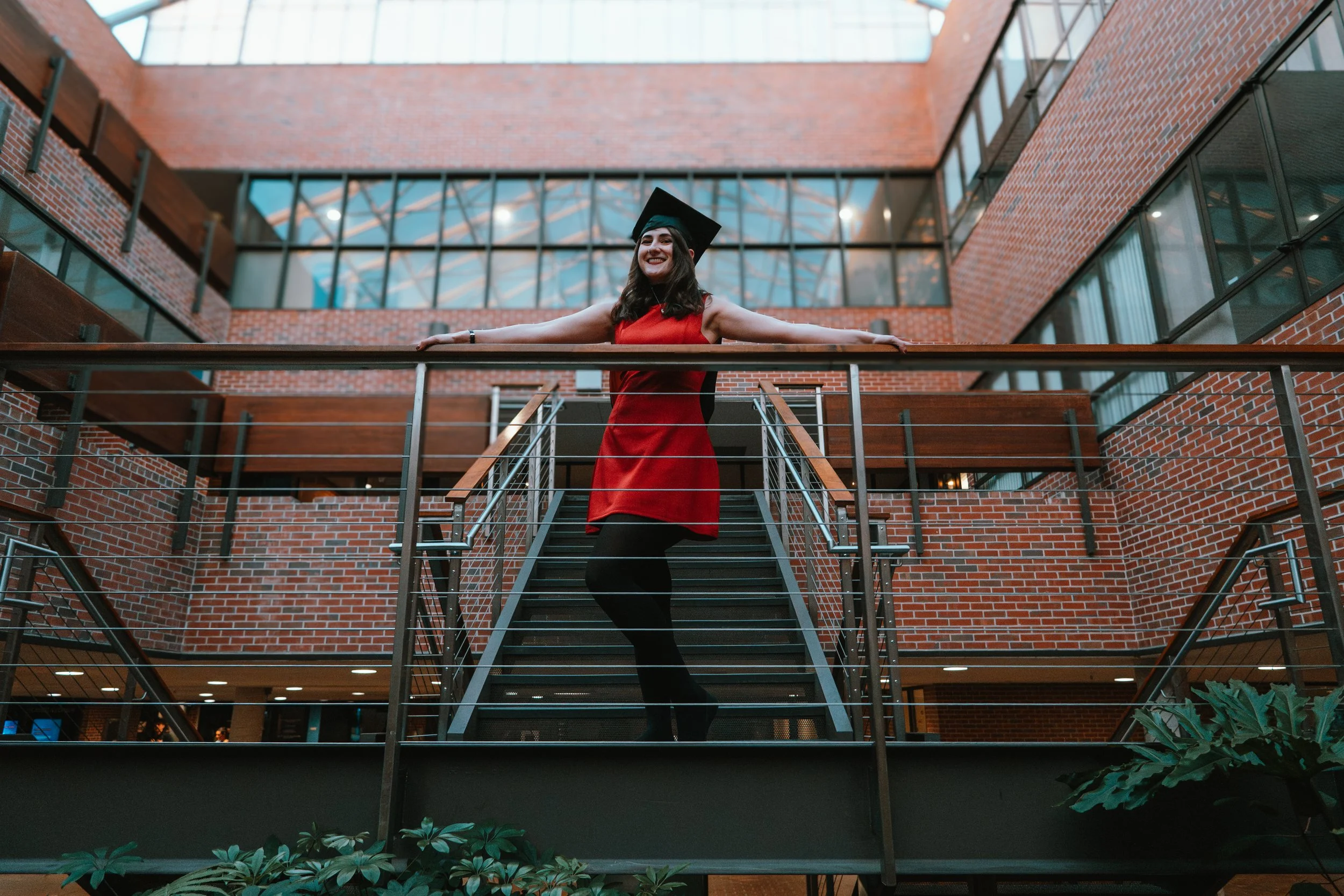 A woman in a red dress and black tights stands on a staircase inside a modern brick building, wearing a graduation cap and smiling with her arms outstretched.