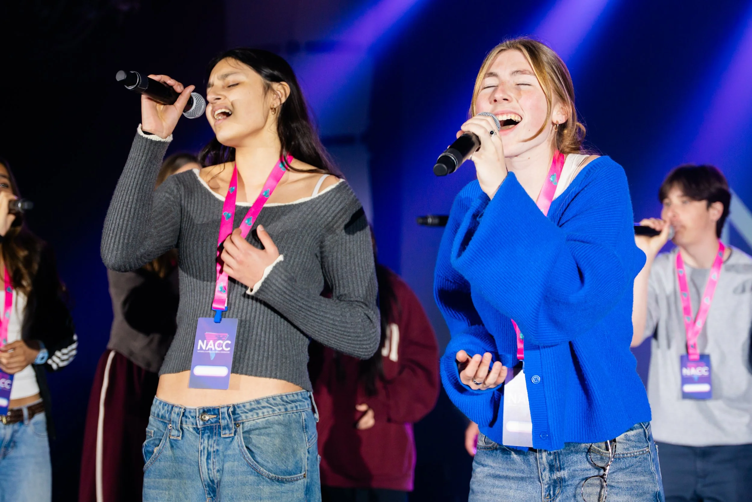 Two young women are singing into microphones on stage at an event, with others visible in the background. One has dark hair and is wearing a gray sweater and jeans, the other has light hair and is dressed in a blue jacket and jeans. Both are wearing 