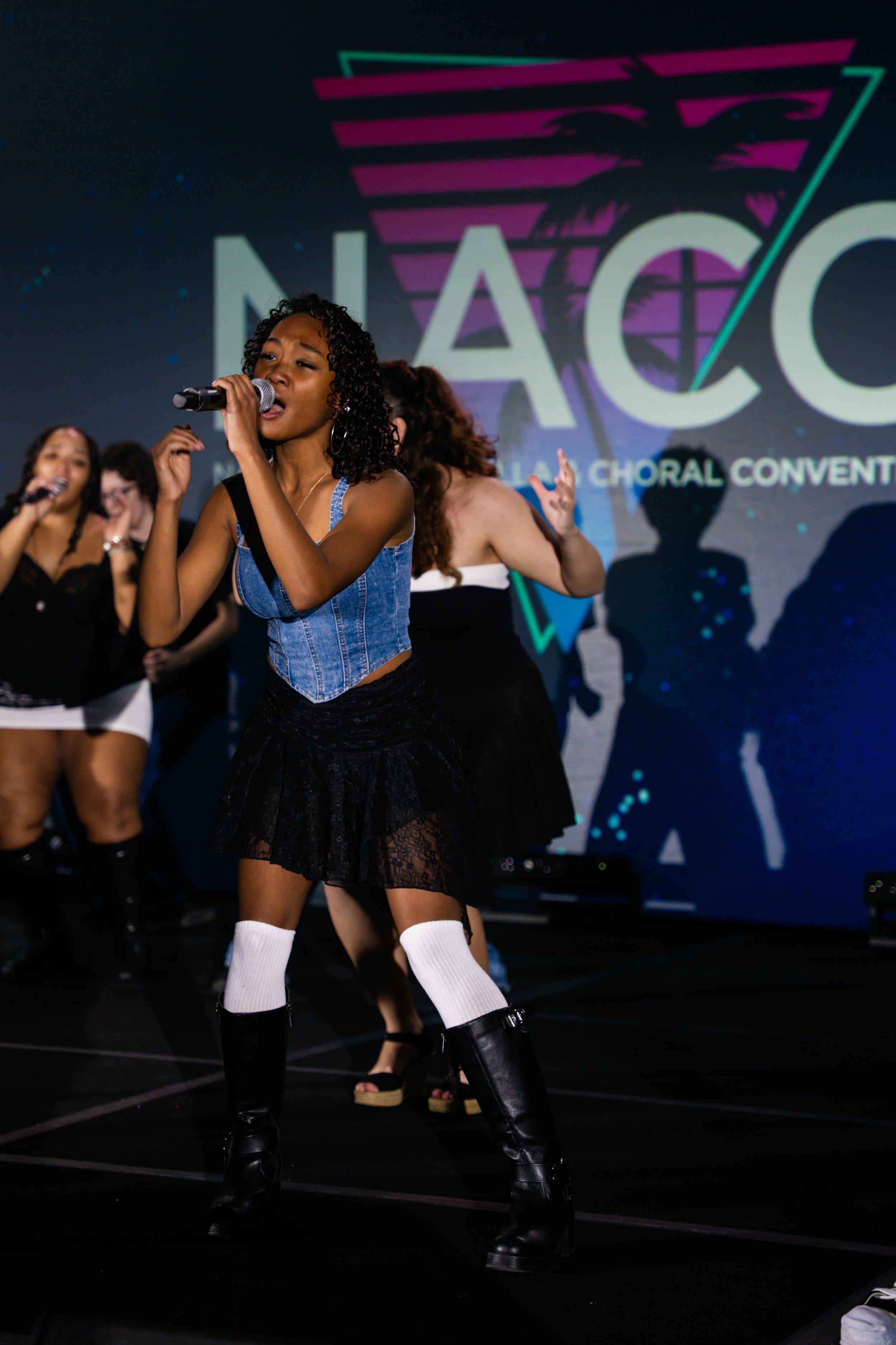 Young woman with curly hair singing into a microphone on stage during a choir performance at the NACC event, with other choir members in the background and a large colorful logo projected on the wall behind her.