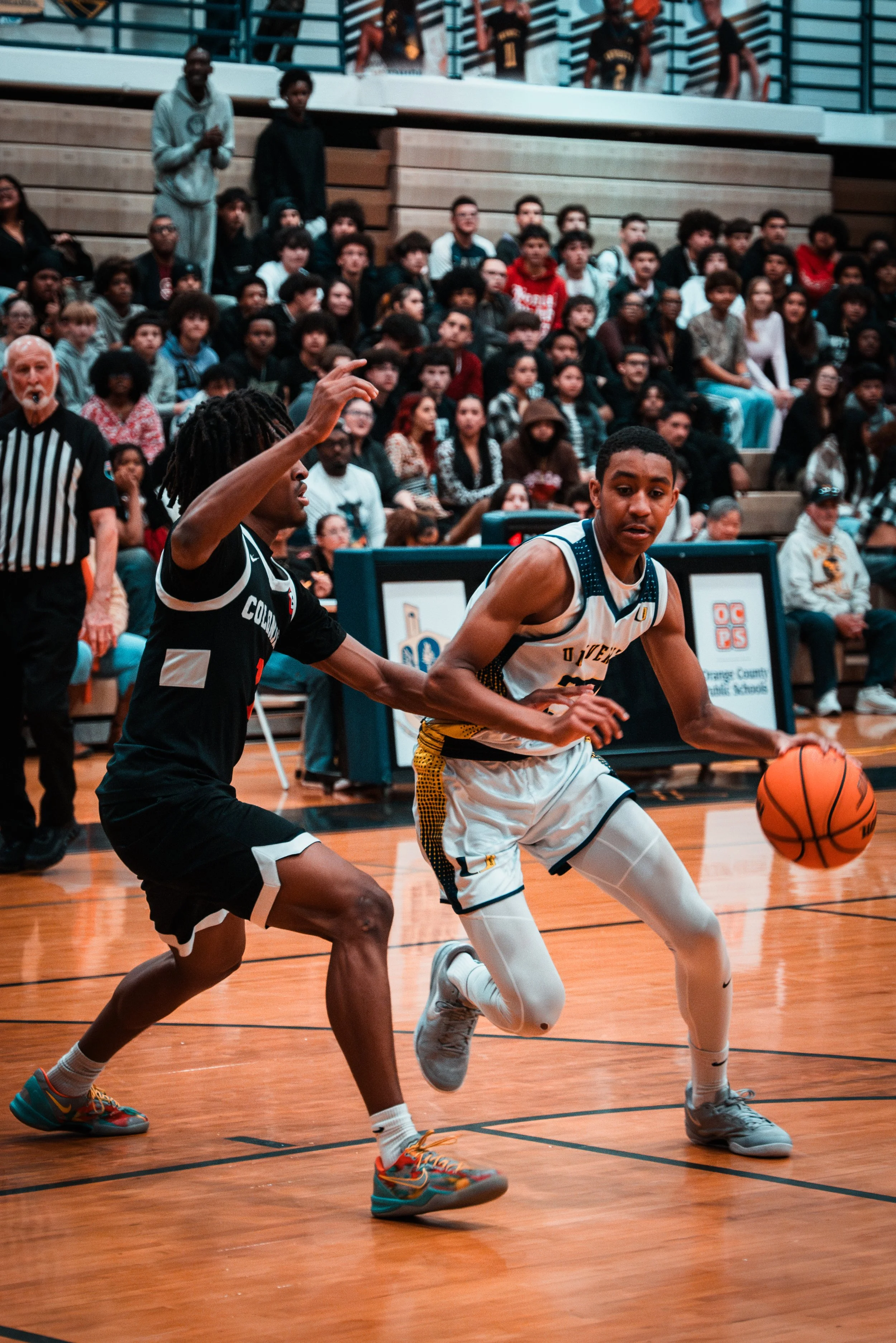Two young basketball players compete for the ball on a gym court, with a large audience of spectators seated behind them.