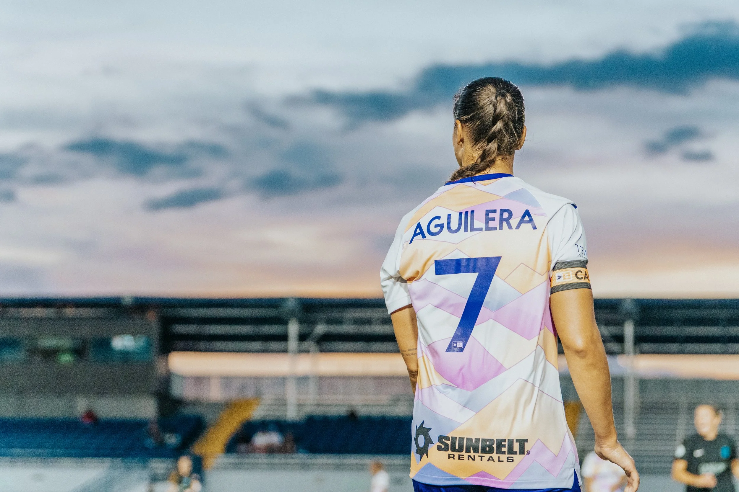 Back of a female soccer player with a braid, wearing a colorful jersey with the name 'Aguilera' and the number 7, standing on a soccer field during sunset.