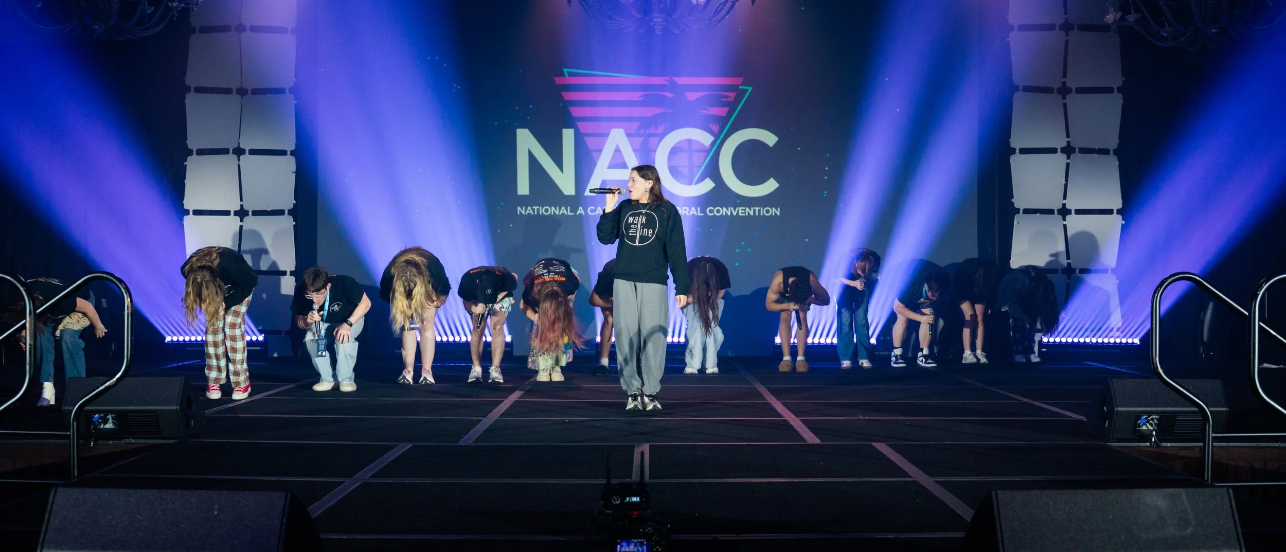 A group of children and a woman performing on a stage during the National A Capella Conference (NACC). The woman is holding a microphone, and the children are bowing.