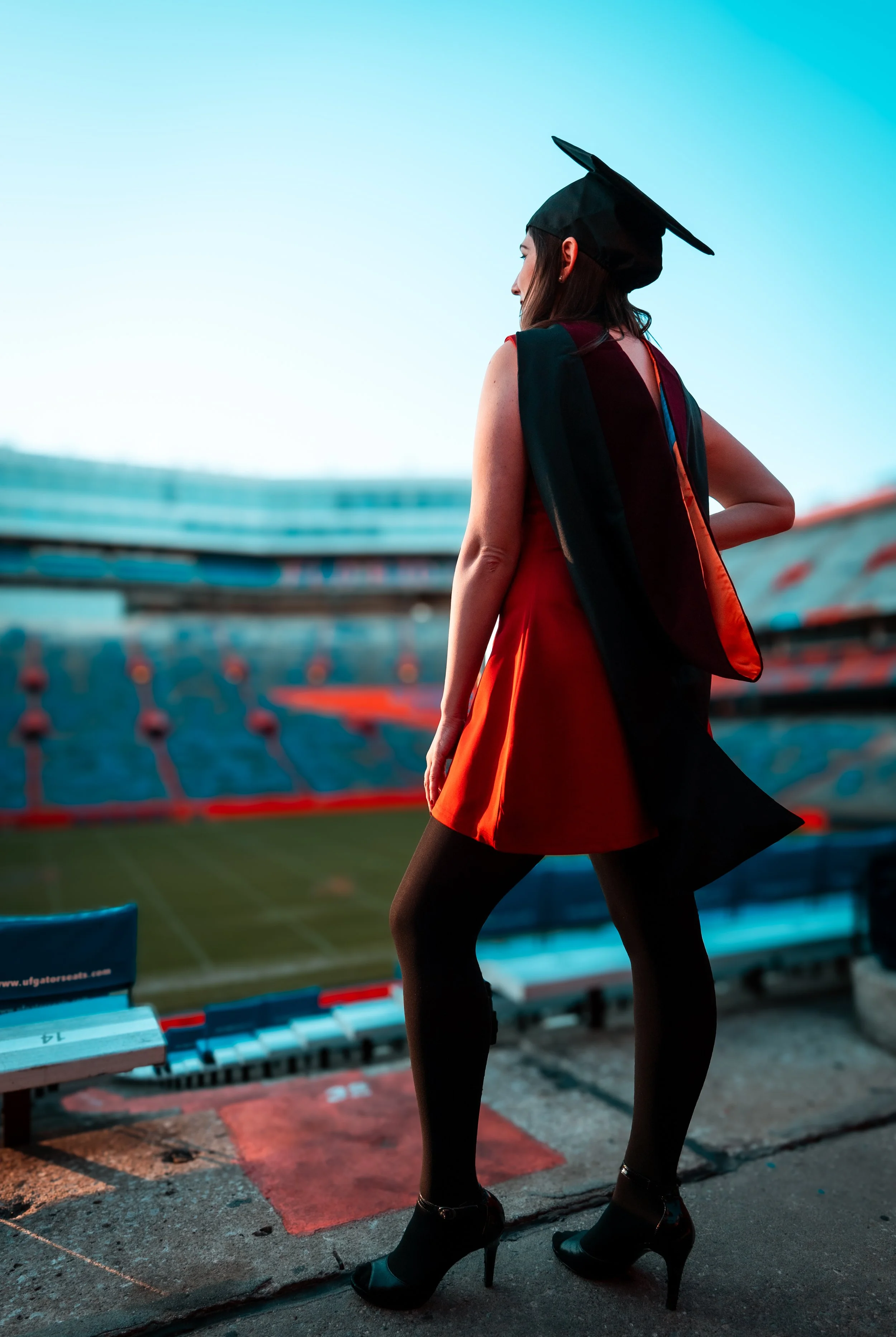 A woman in a red dress and black high heels standing in a stadium, wearing a graduation cap and gown, looking over the field during sunset.