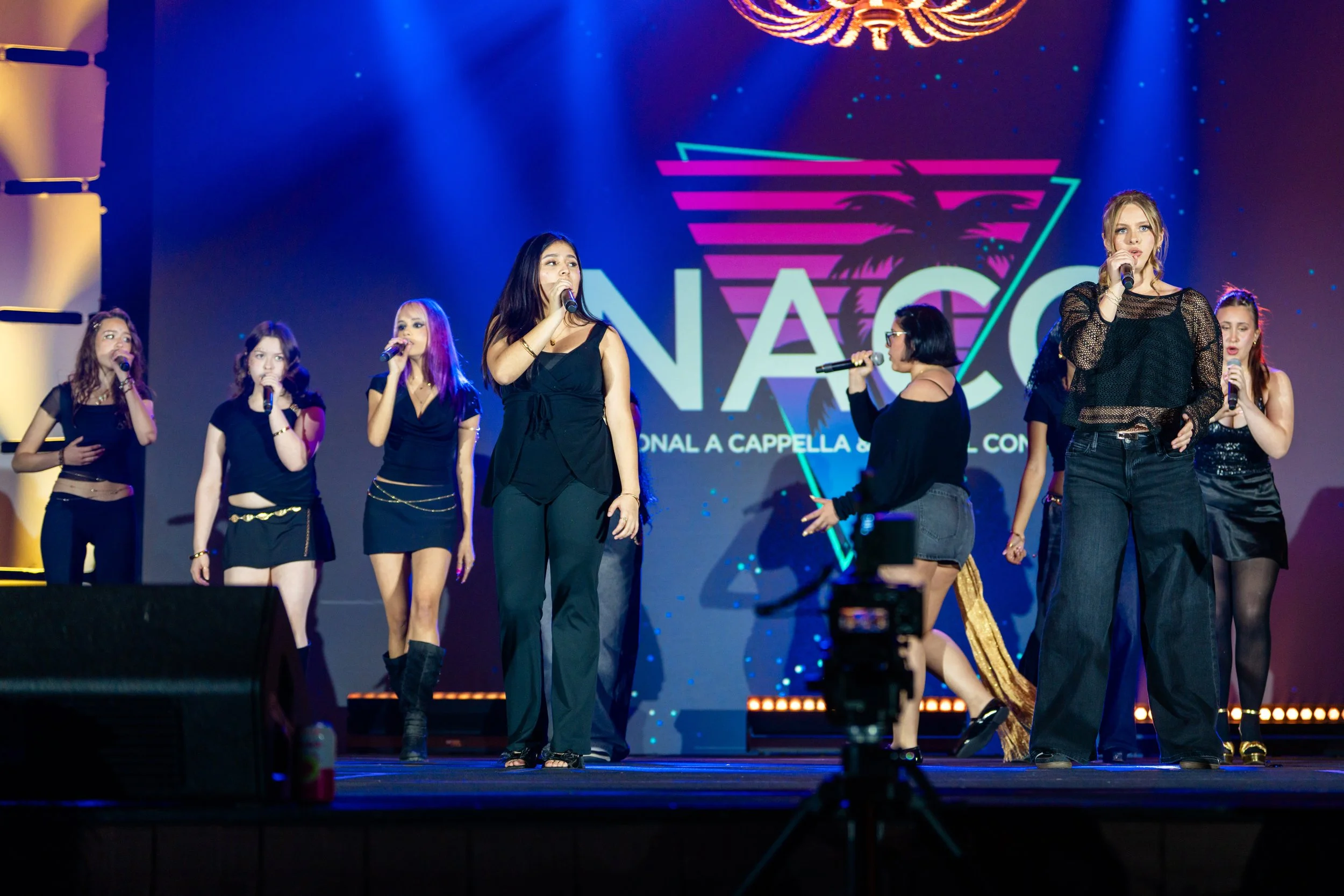 Group of women singing and performing on stage at an event, with a large screen behind them displaying the logo NACO and text about a cappella concert.