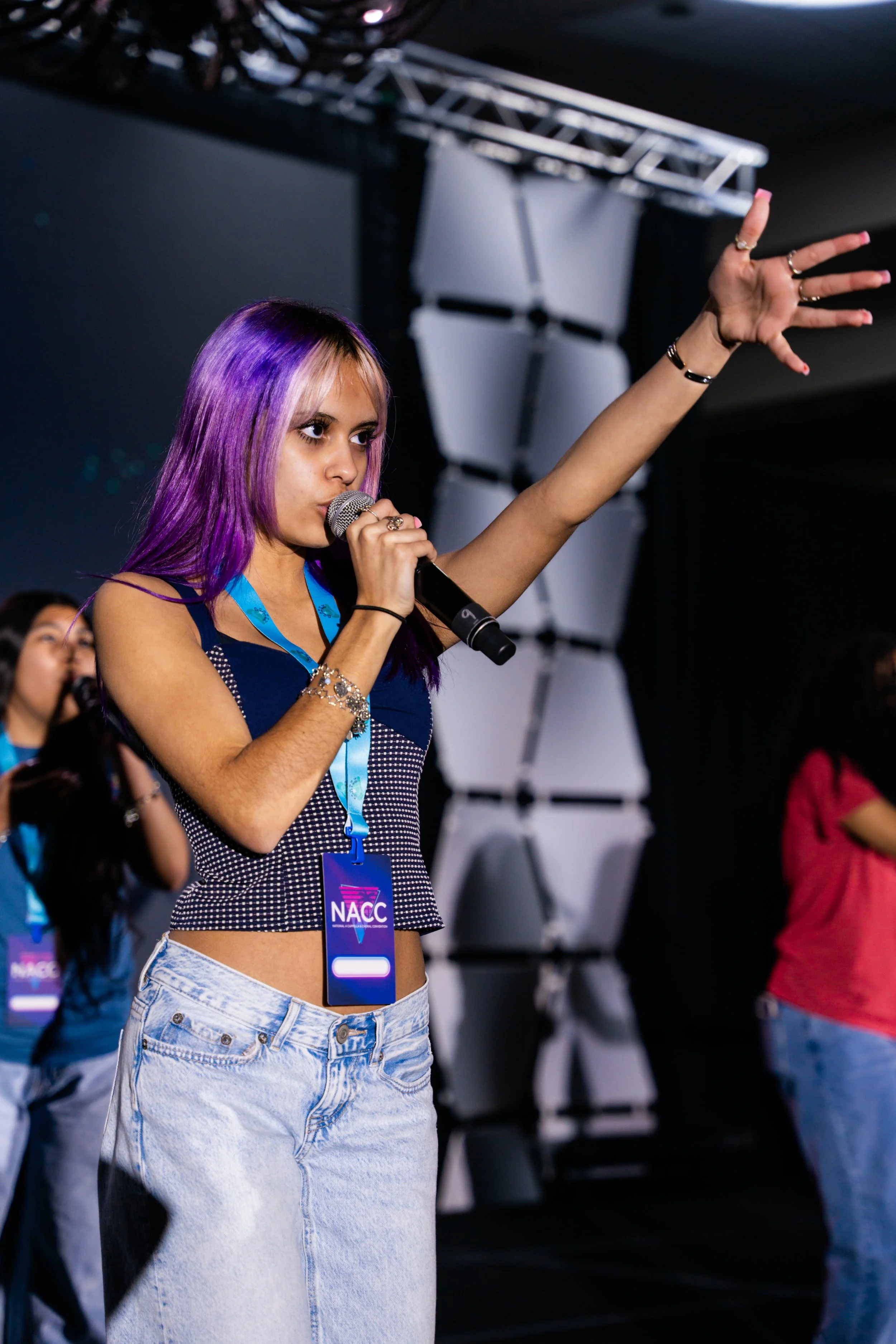 A young woman with purple and blonde hair holding a microphone and wearing a black and white checkered crop top, light blue jeans, and multiple rings and bracelets, at an event with a North American Culture Center (NACC) badge around her neck, with o