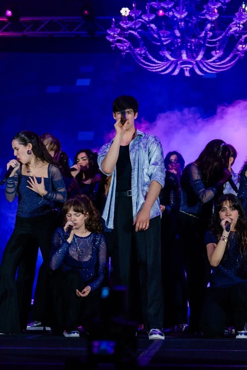 Teenagers singing on stage during a performance with purple lighting and a chandelier overhead.