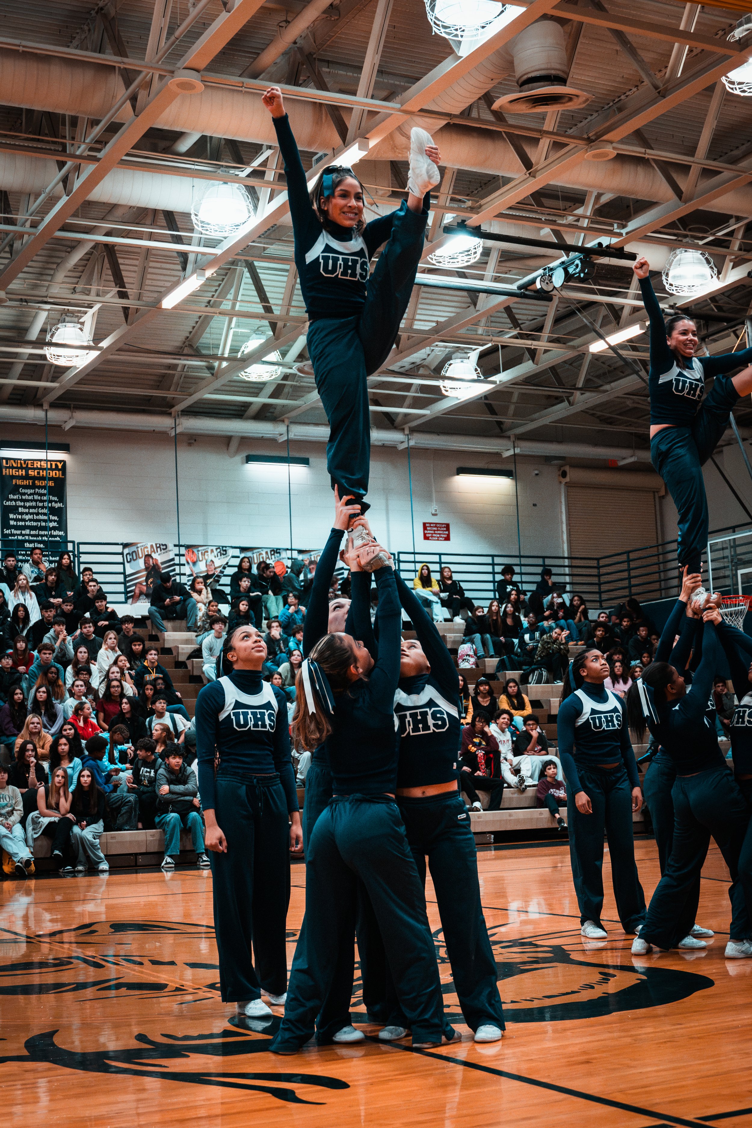 Cheerleaders performing a stunt during a pep rally or game in a gymnasium, with a large audience seated in the background.