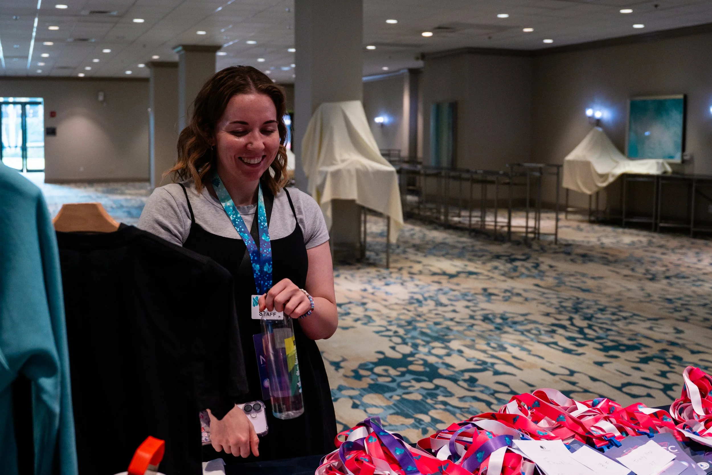 A smiling woman with brown hair, wearing a staff badge, standing at a table filled with colorful ribbons and papers in a conference or event hall.