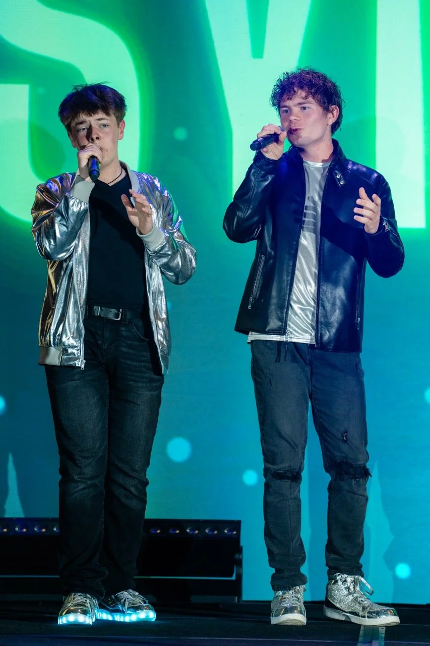 Two young men singing into microphones on stage with a green and blue digital background.