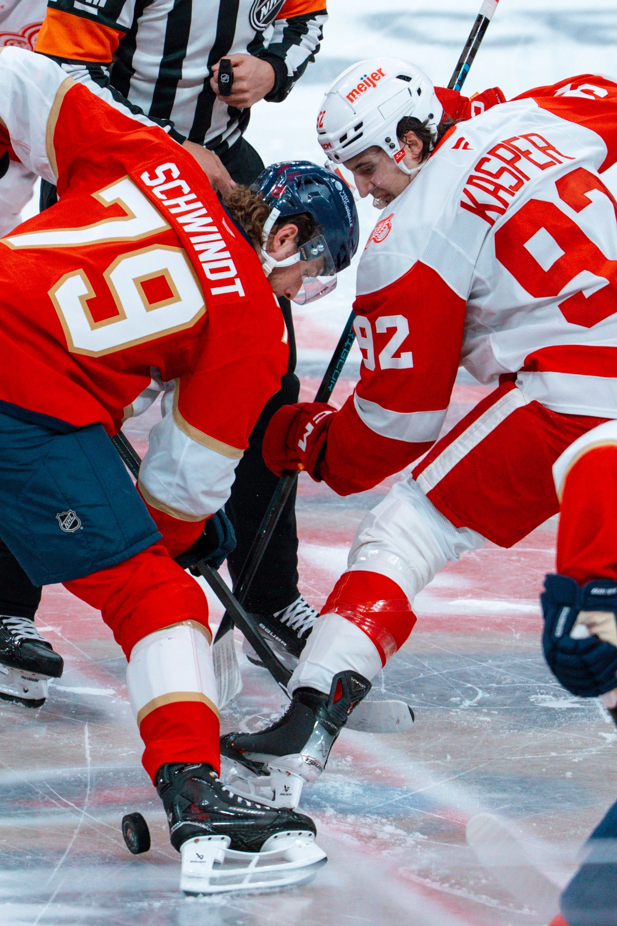Hockey players face off during a game on ice, wearing protective gear and uniforms, with a referee overseeing.