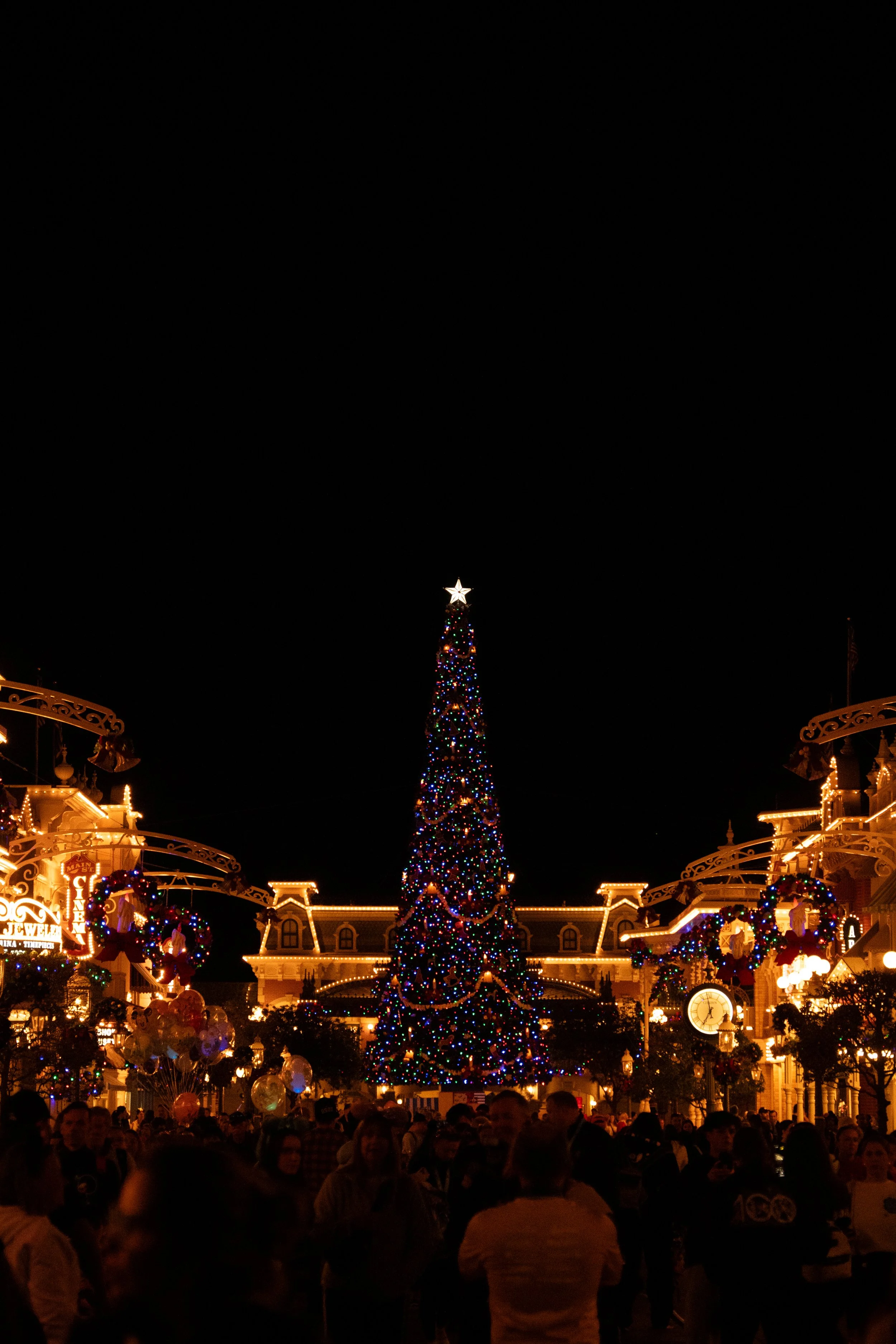 Nighttime scene at a theme park with a large, decorated Christmas tree in the center, surrounded by people, colorful lights, and festive wreaths.