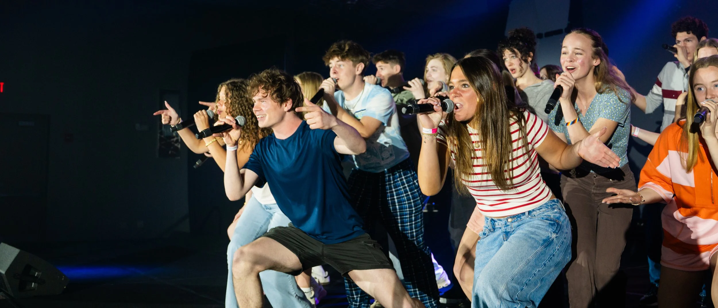 Group of young people singing and dancing on stage with microphones, wearing casual clothes, having fun.