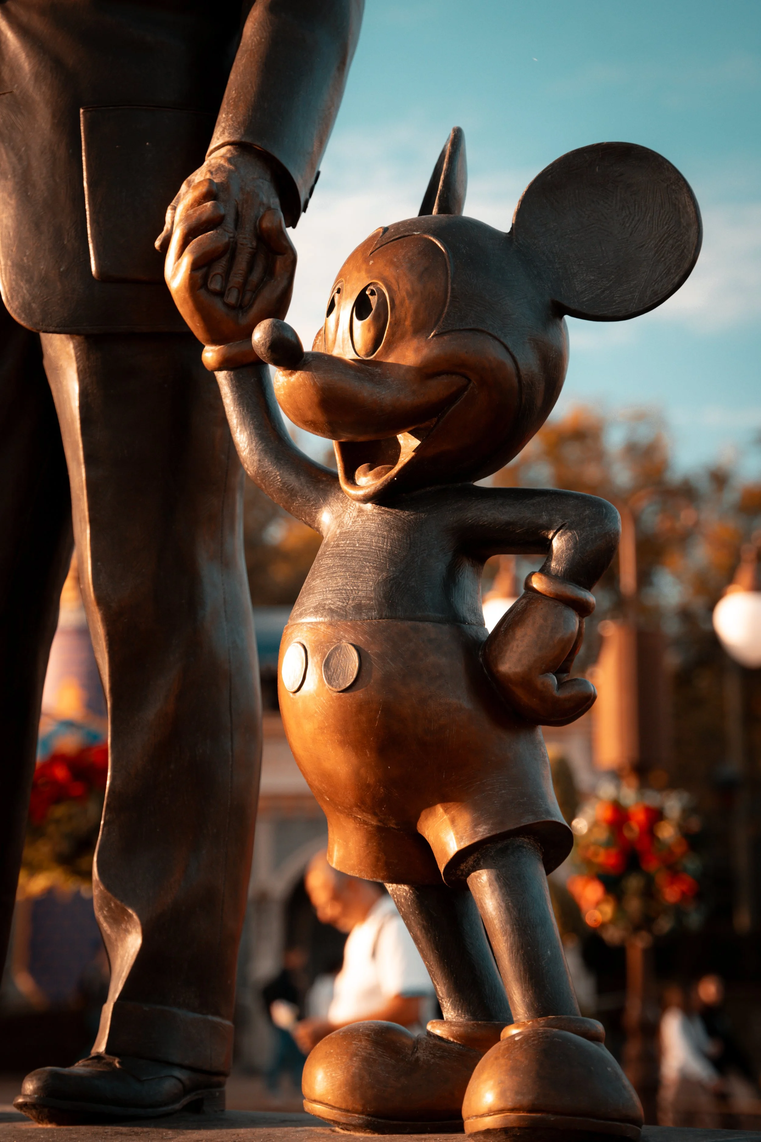 A bronze statue of Mickey Mouse holding hands with a man in a suit, with a sunset background and people in the distance.