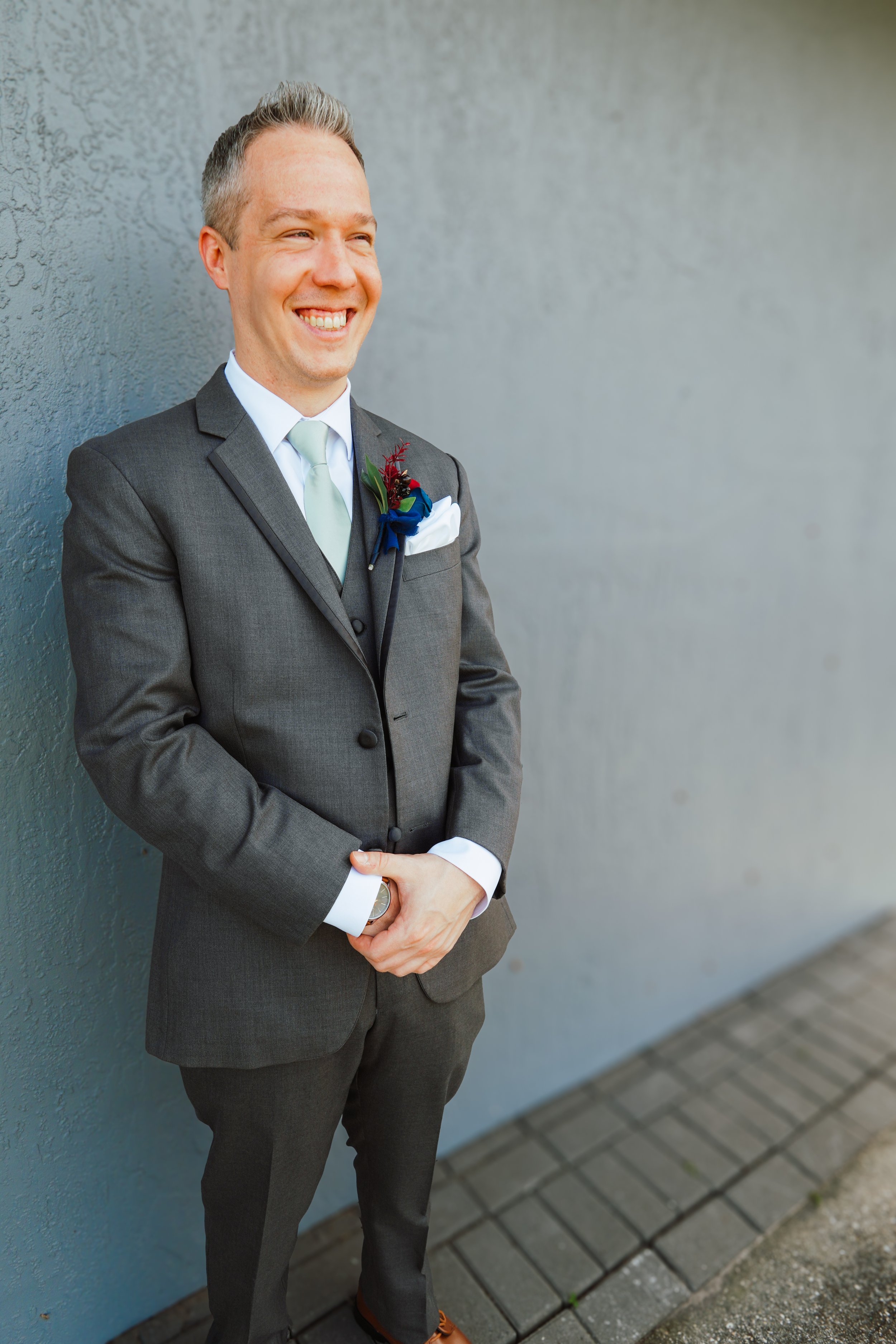 A smiling man in a gray suit with a boutonniere, standing against a gray wall outside.