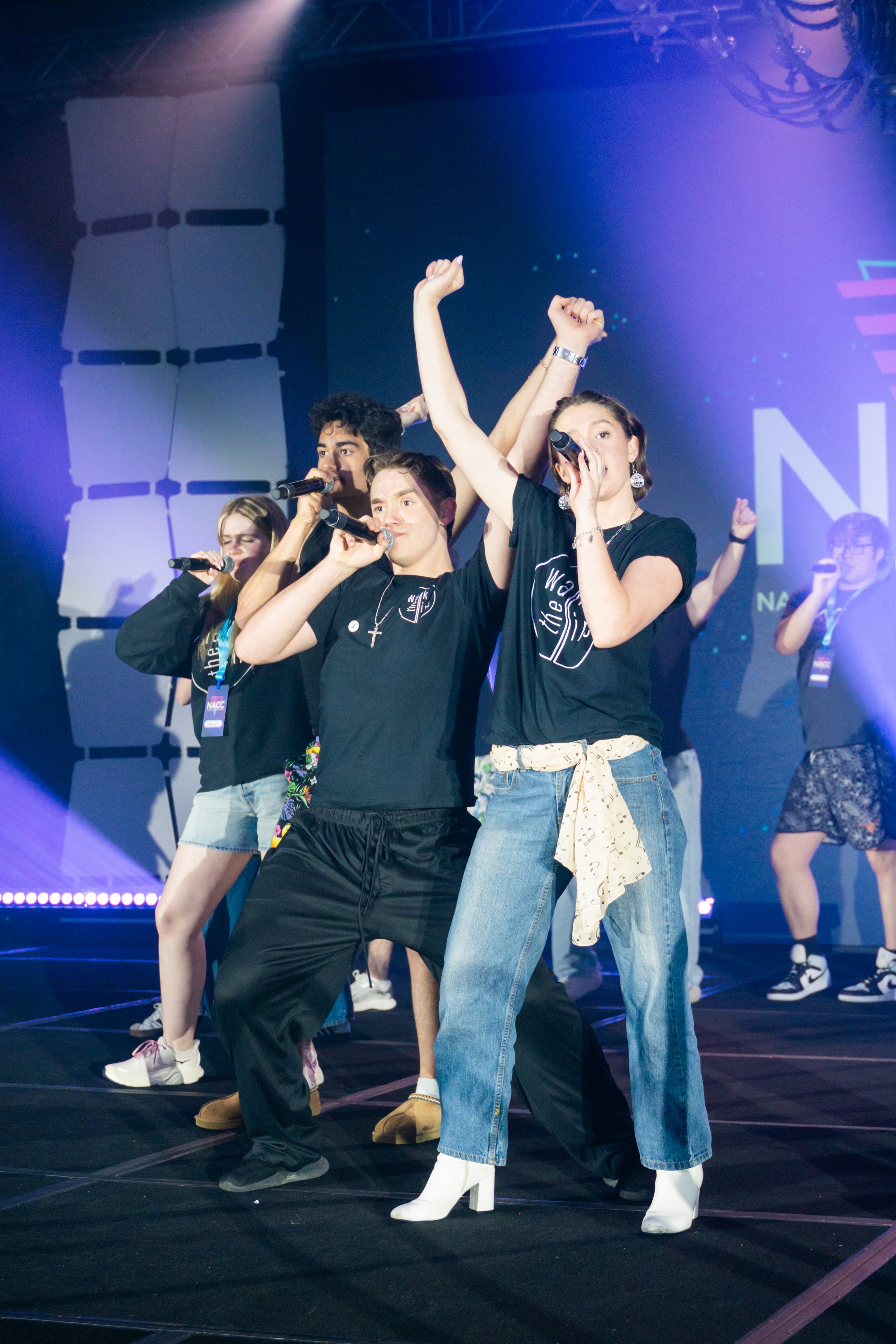 A group of young people performing on stage, singing into microphones, with some raising their fists and striking poses. The backdrop displays a large screen with colorful graphics and text.