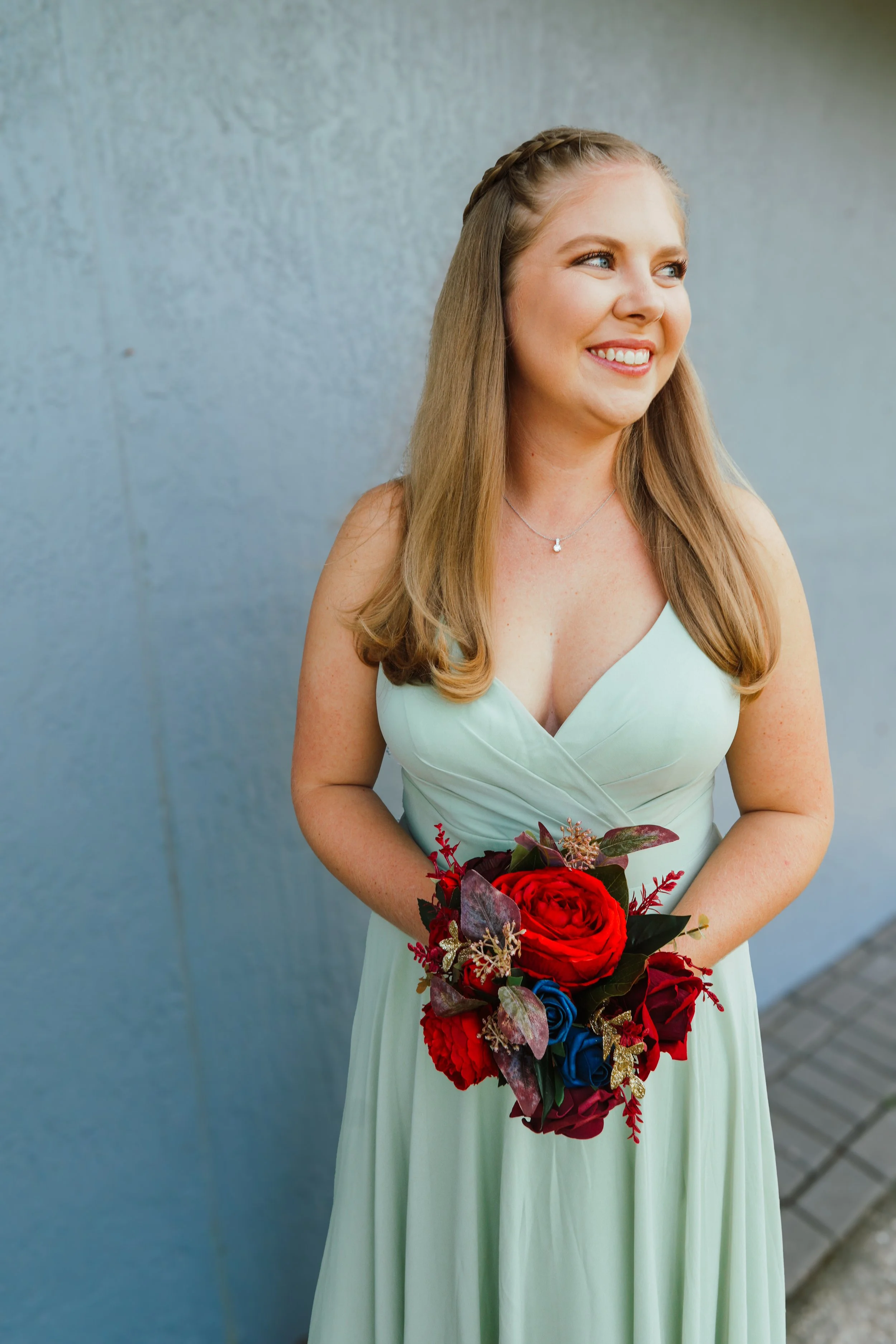 A smiling woman in a light green dress holding a bouquet of red, blue, and purple flowers, standing against a gray wall.