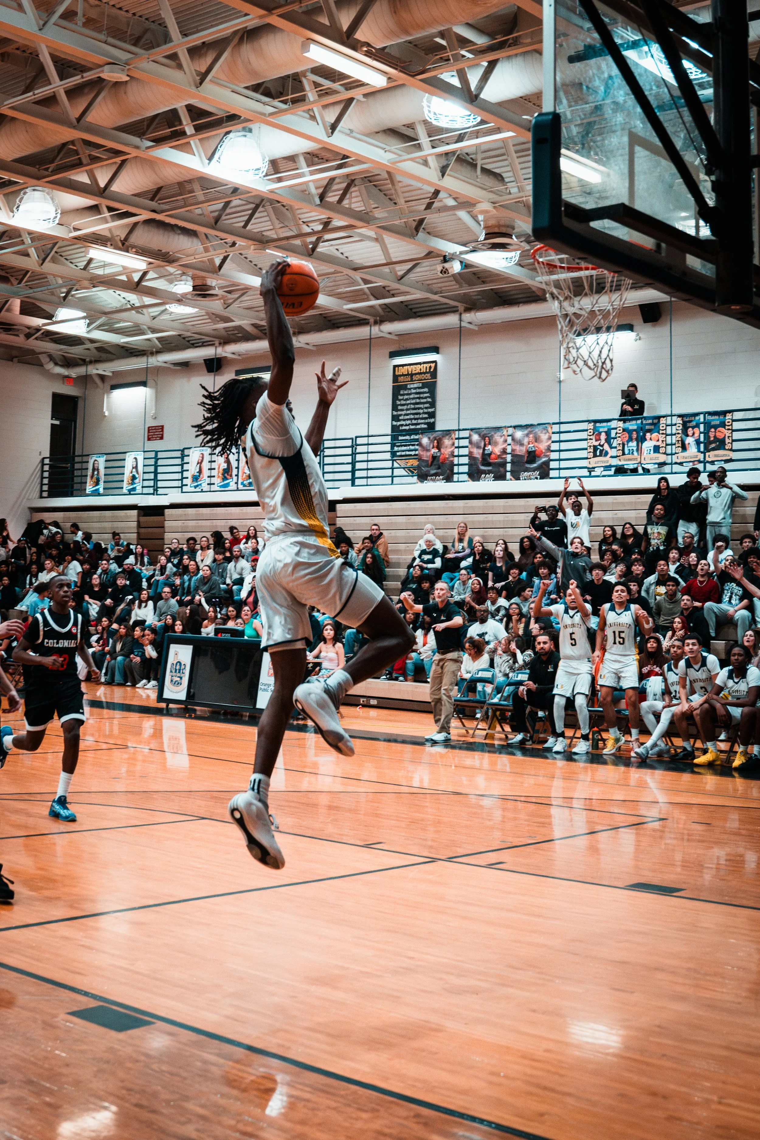 A basketball player in a white uniform jumps to make a shot during a game, with a crowd watching from the bleachers and teammates on the sideline.