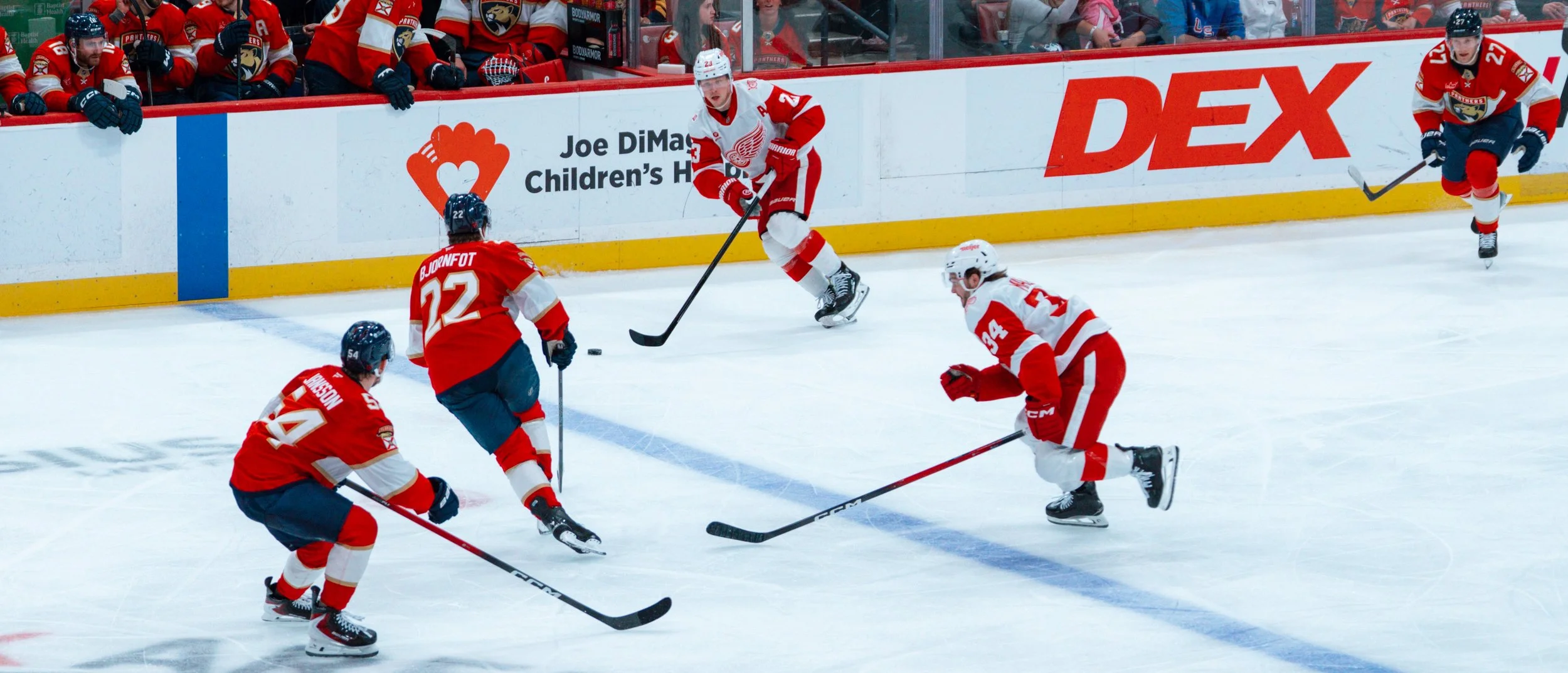 Hockey players in red and white uniforms competing during a game on the ice rink with spectators and advertising banners in the background.