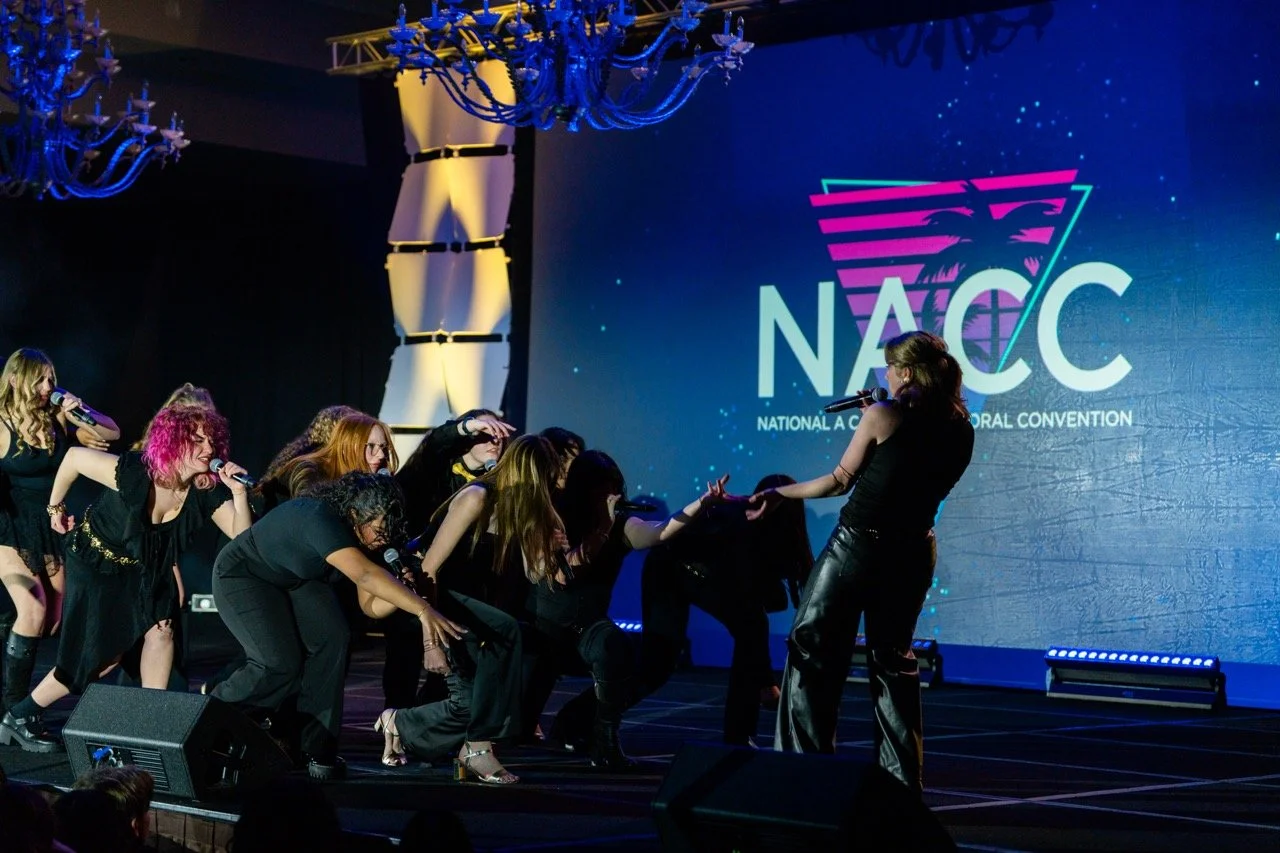 A group of women are performing on stage at the National A Cappella Conference, with one woman singing into a microphone and others dancing and singing. The background displays the NABCC logo.