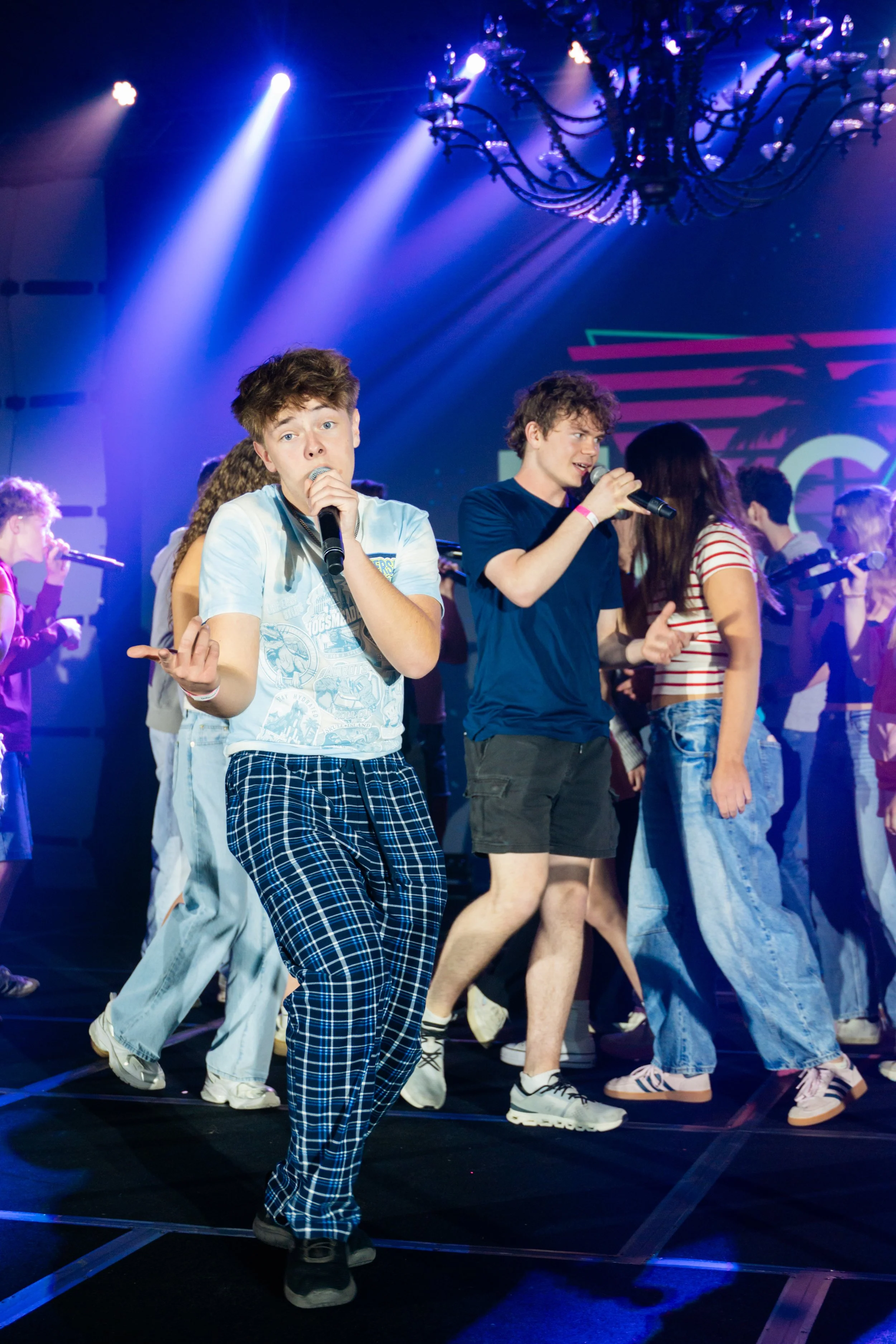 Group of young performers on stage participating in a dance or musical performance, with colorful stage lighting and a chandelier overhead.