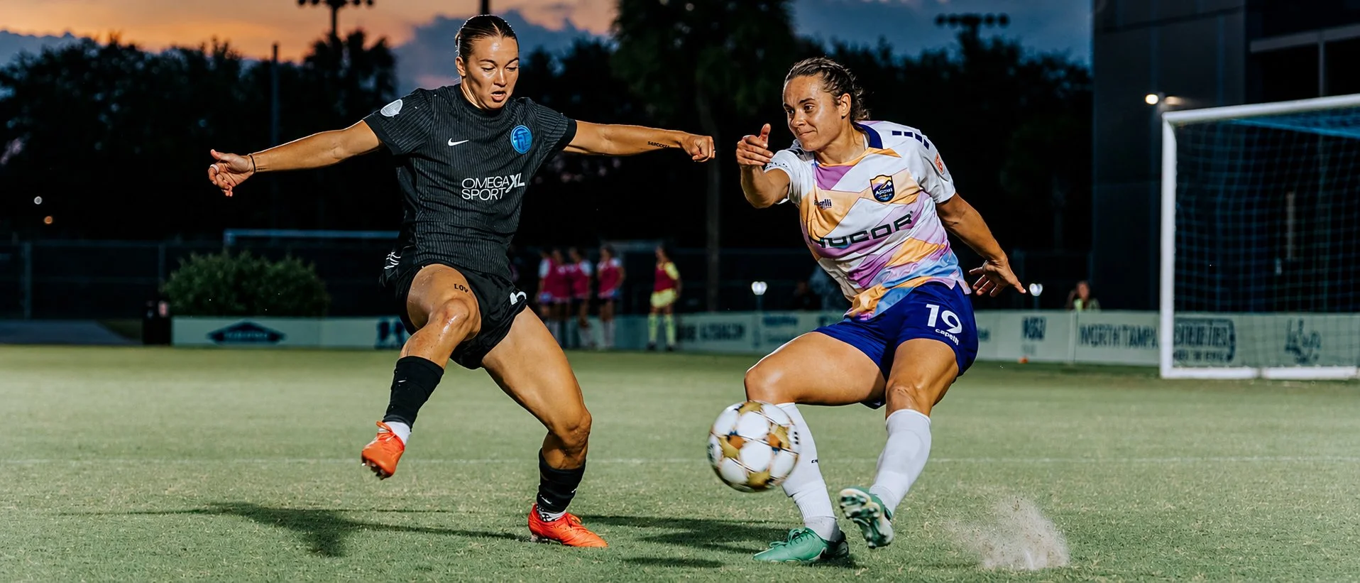 Two female soccer players competing for the ball on a field during sunset, with a goal post in the background.