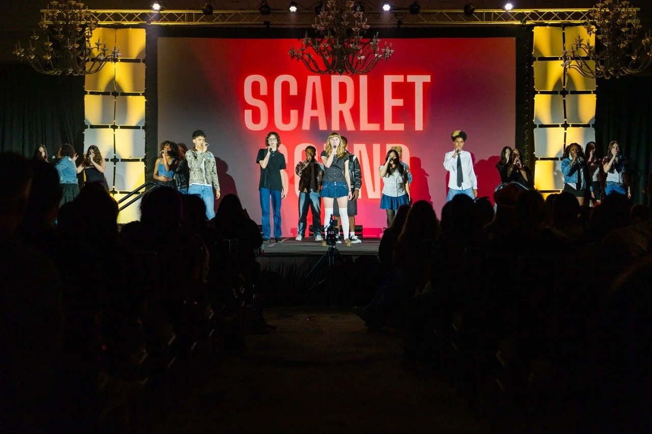 Students perform on stage at a school event with a large screen displaying 'SCARLET SQUAD' in the background, audience watching in the dark.