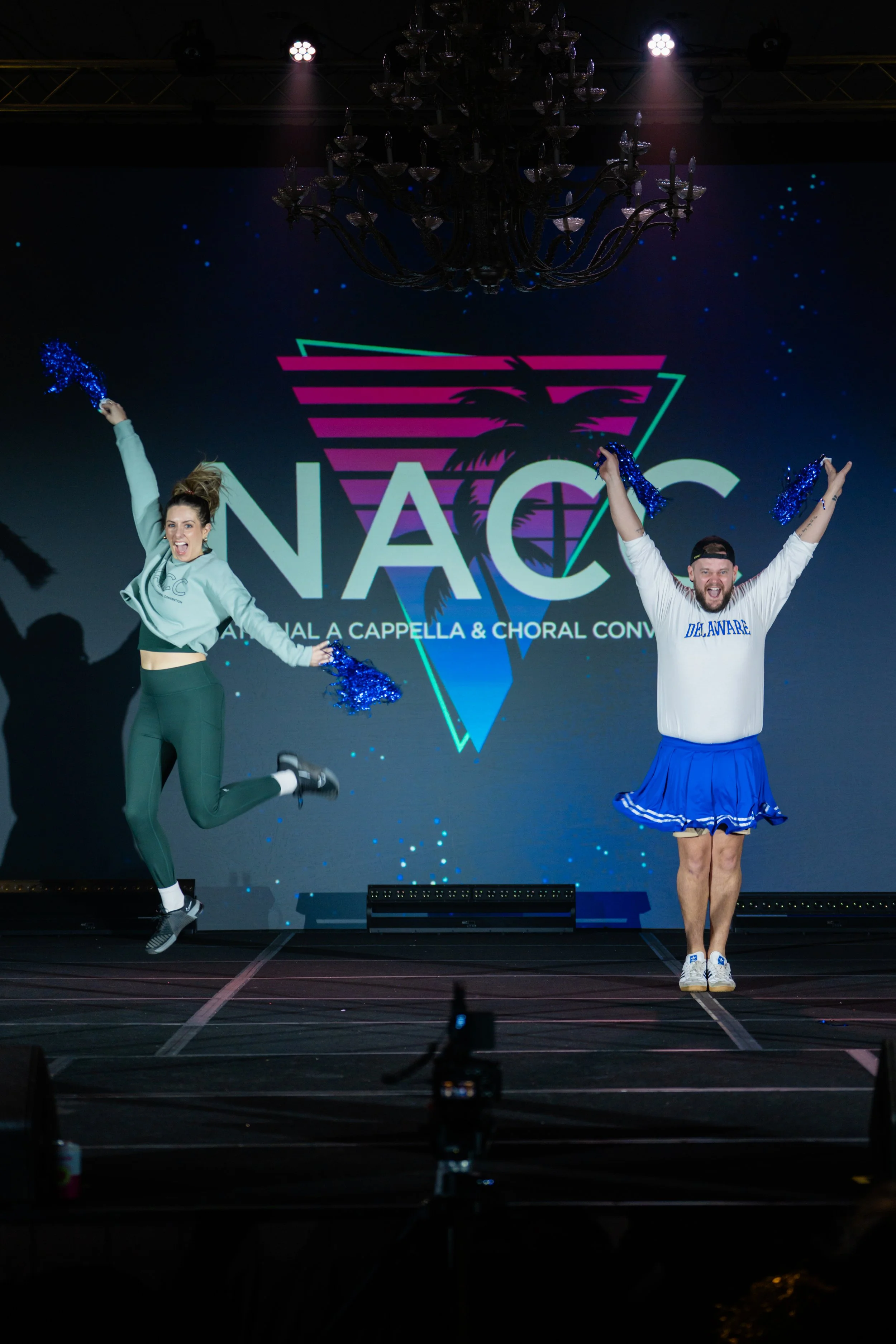 Two performers on stage under bright spotlights, jumping and smiling, with a large LED screen behind them displaying a logo for the NAC (National A Cappella & Choral Conference). The woman on the left is wearing a light green cropped hoodie and dark 