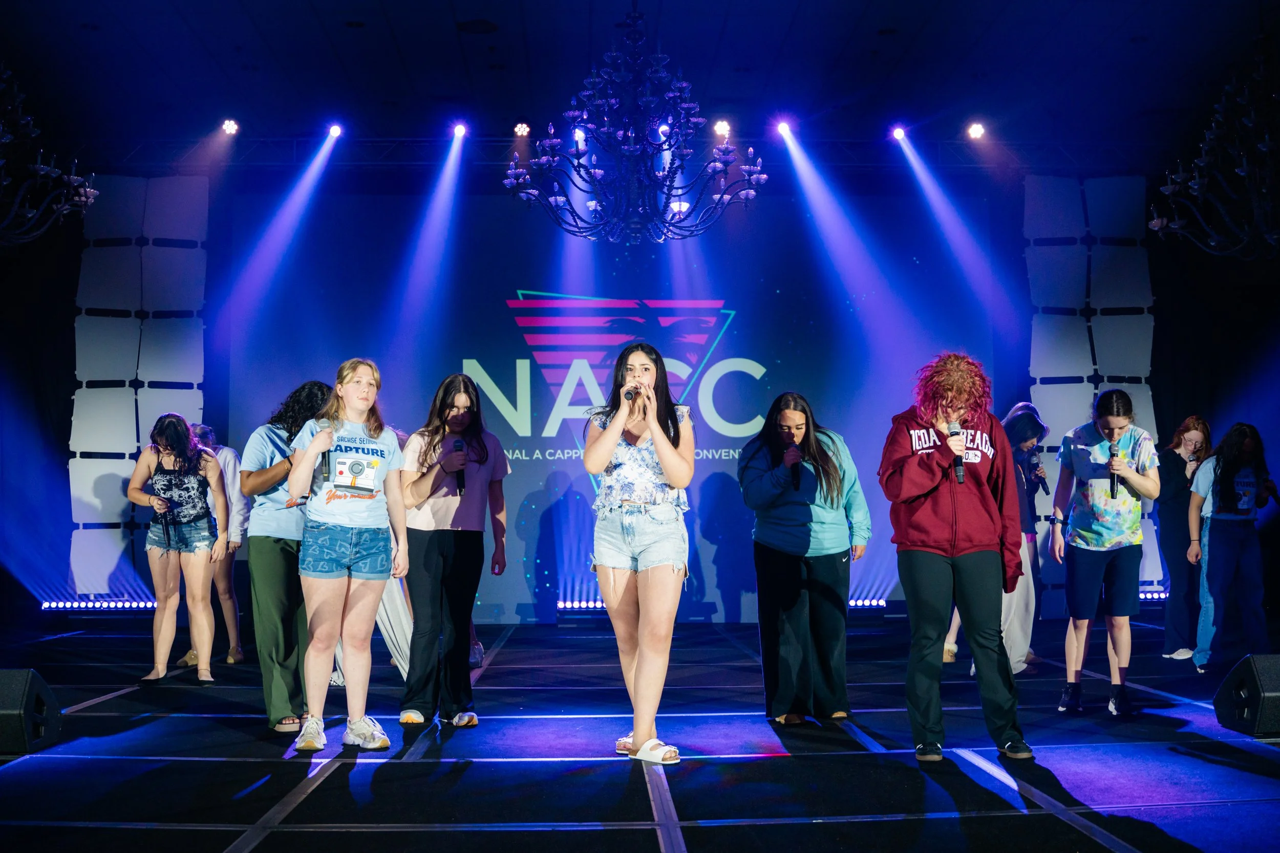 A group of young women standing on a stage with bright colorful lights, some holding microphones. Behind them is a large screen displaying a logo that reads 'NACC.' The stage has a chandelier hanging from the ceiling.