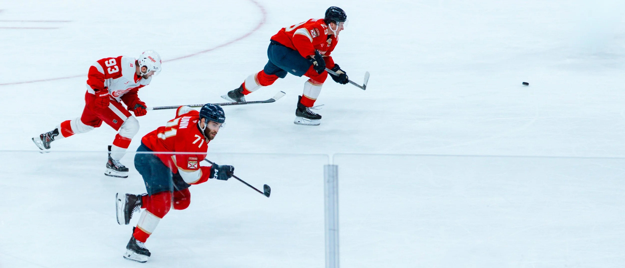 Three hockey players in red uniforms are skating quickly on ice, chasing after a puck during a game.