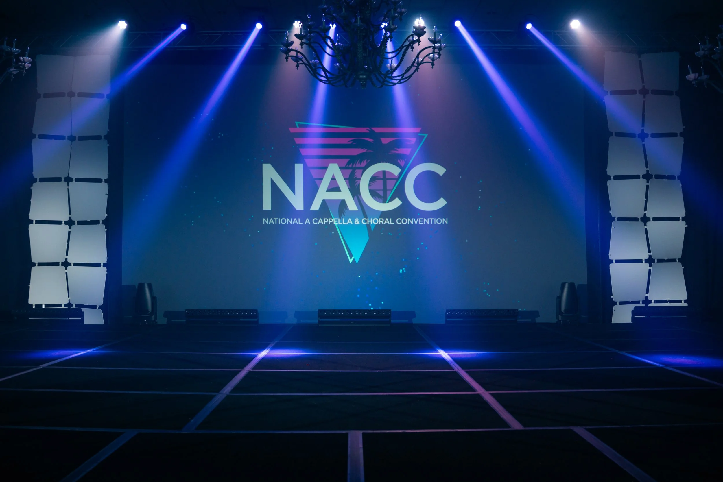 Stage with large screen displaying NAC and logo for National A Cappella & Choral Convention, illuminated by blue and purple stage lights.