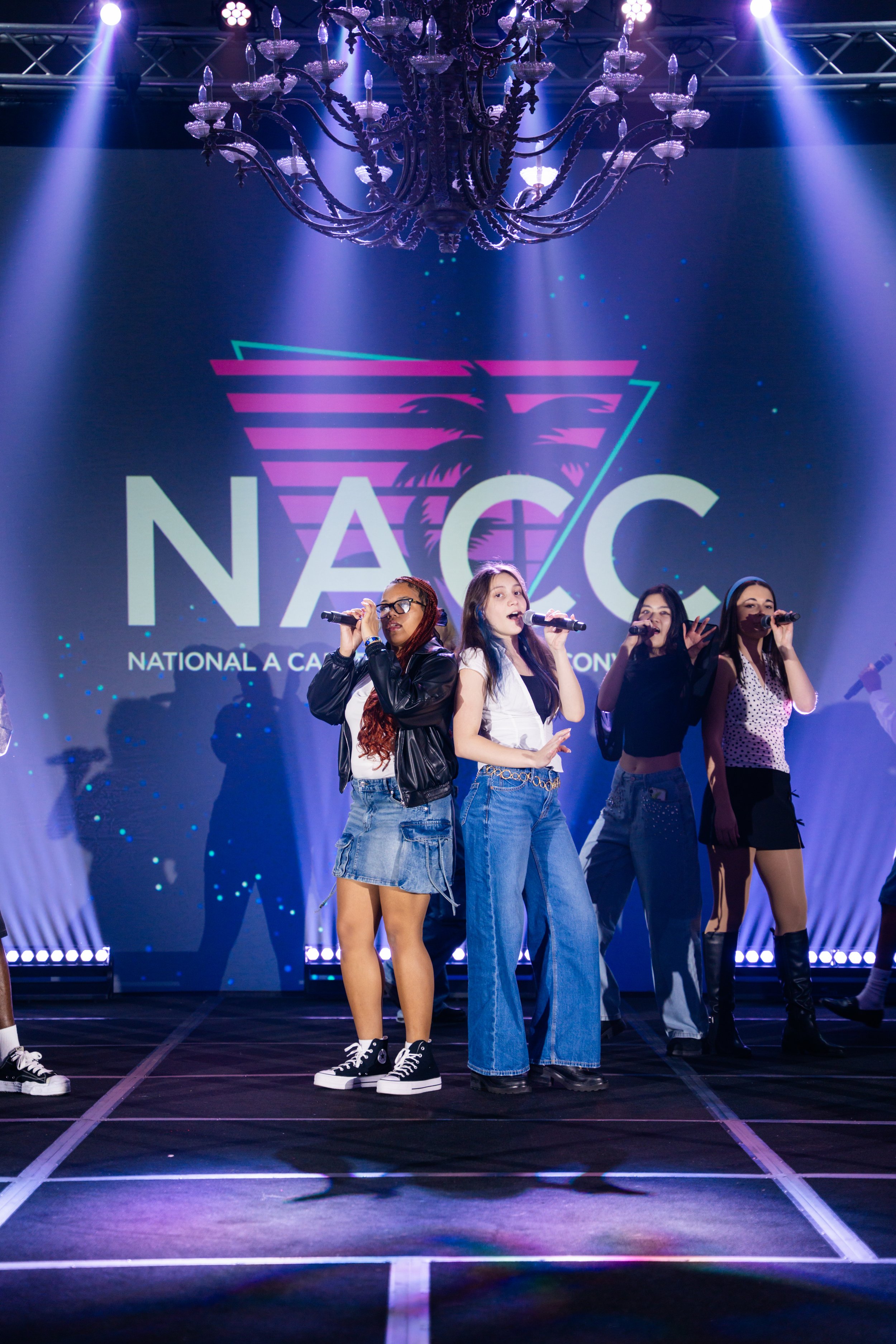 A group of five young women singing on stage with a large screen behind them displaying a logo for N.A.G.C. and the words 'National A Capella Competition.' The stage is lit with multiple spotlights, and a large chandelier hangs above the stage.