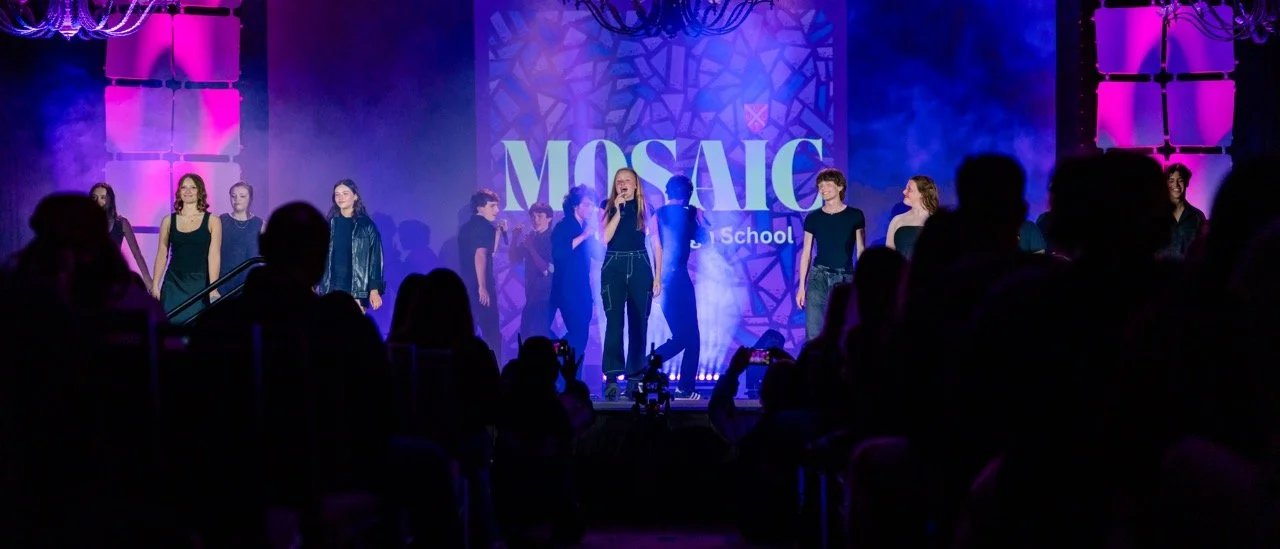 Students performing on stage at Mosaic School event with colorful lighting and audience watching.
