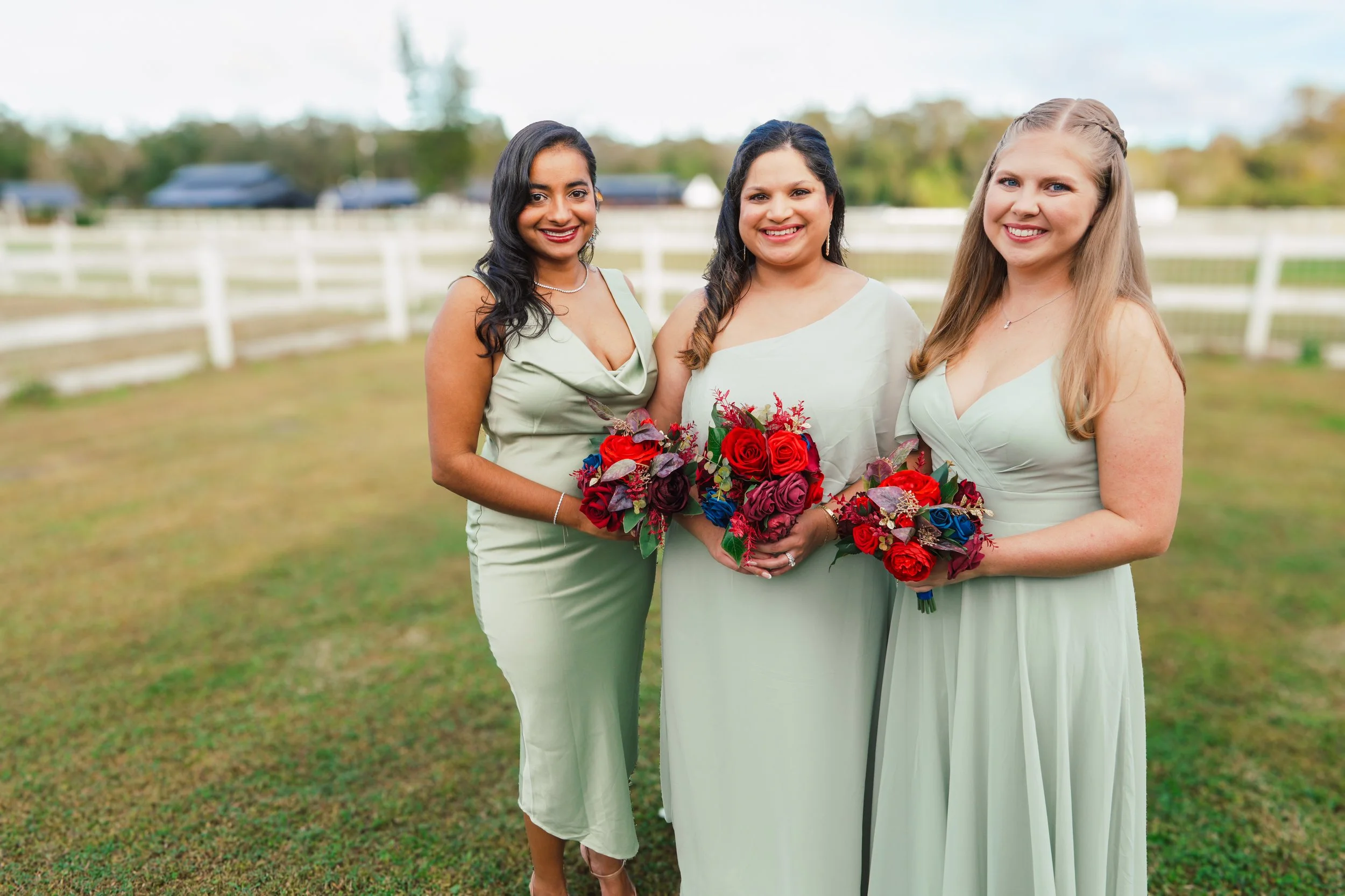 Three women dressed in light green dresses holding bouquets of red, purple, and blue flowers standing outdoors on grass with a white fence and trees in the background.