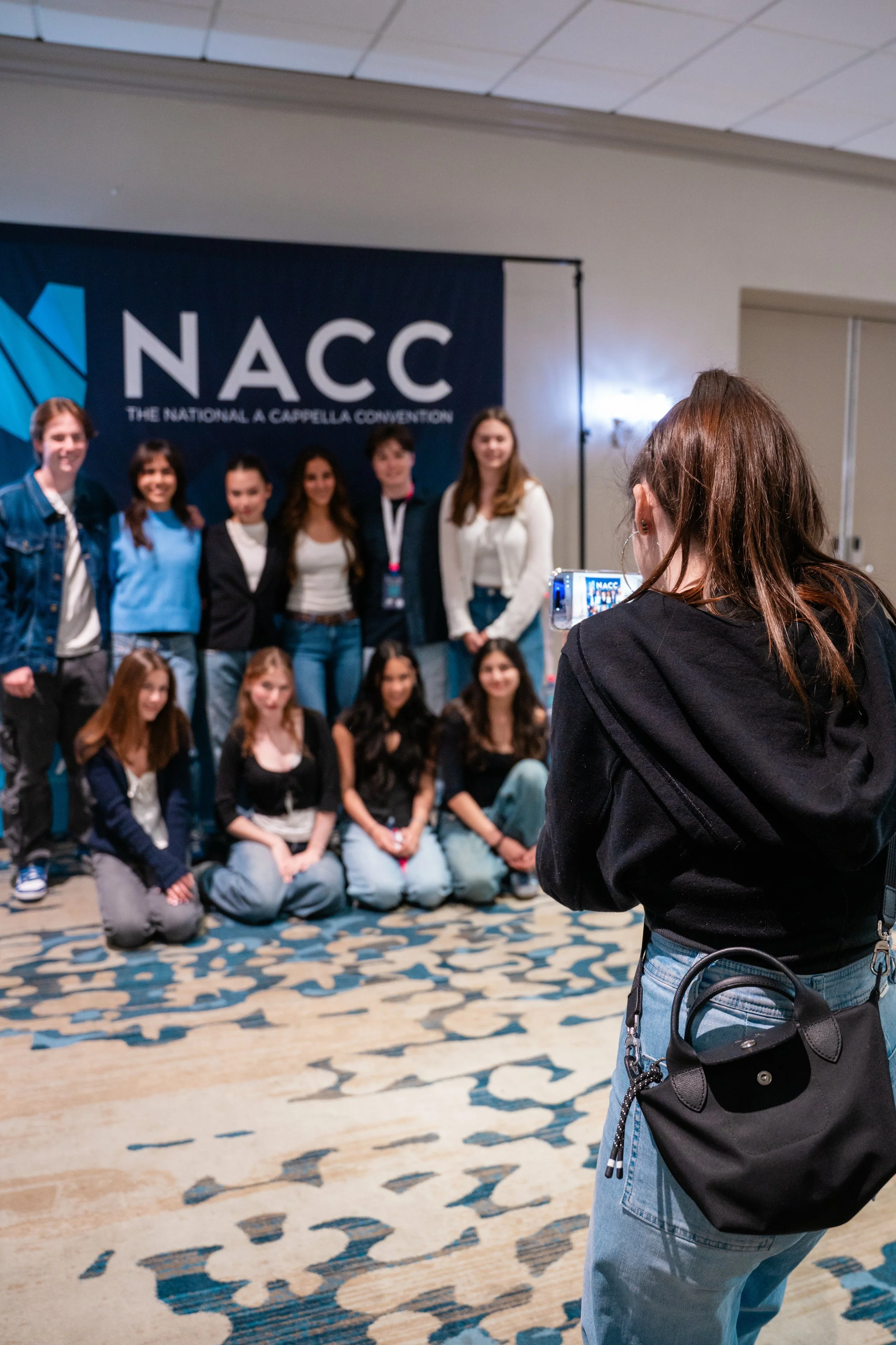 A group of people gathering for a photo at the NACC, the National A Cappella Conference, with a woman in a black jacket taking a picture of them in front of a large blue banner.