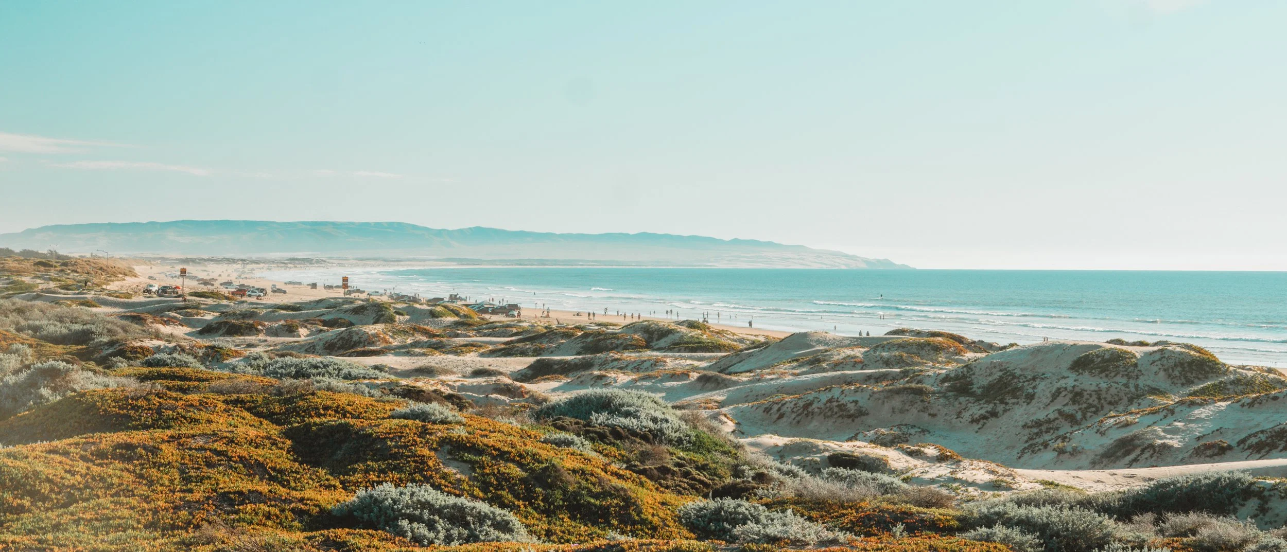 Coastal beach scene with sand dunes covered in low shrubs in the foreground, a sandy beach with people and vehicles in the middle ground, and the ocean with waves and cliffs in the background on a sunny day.