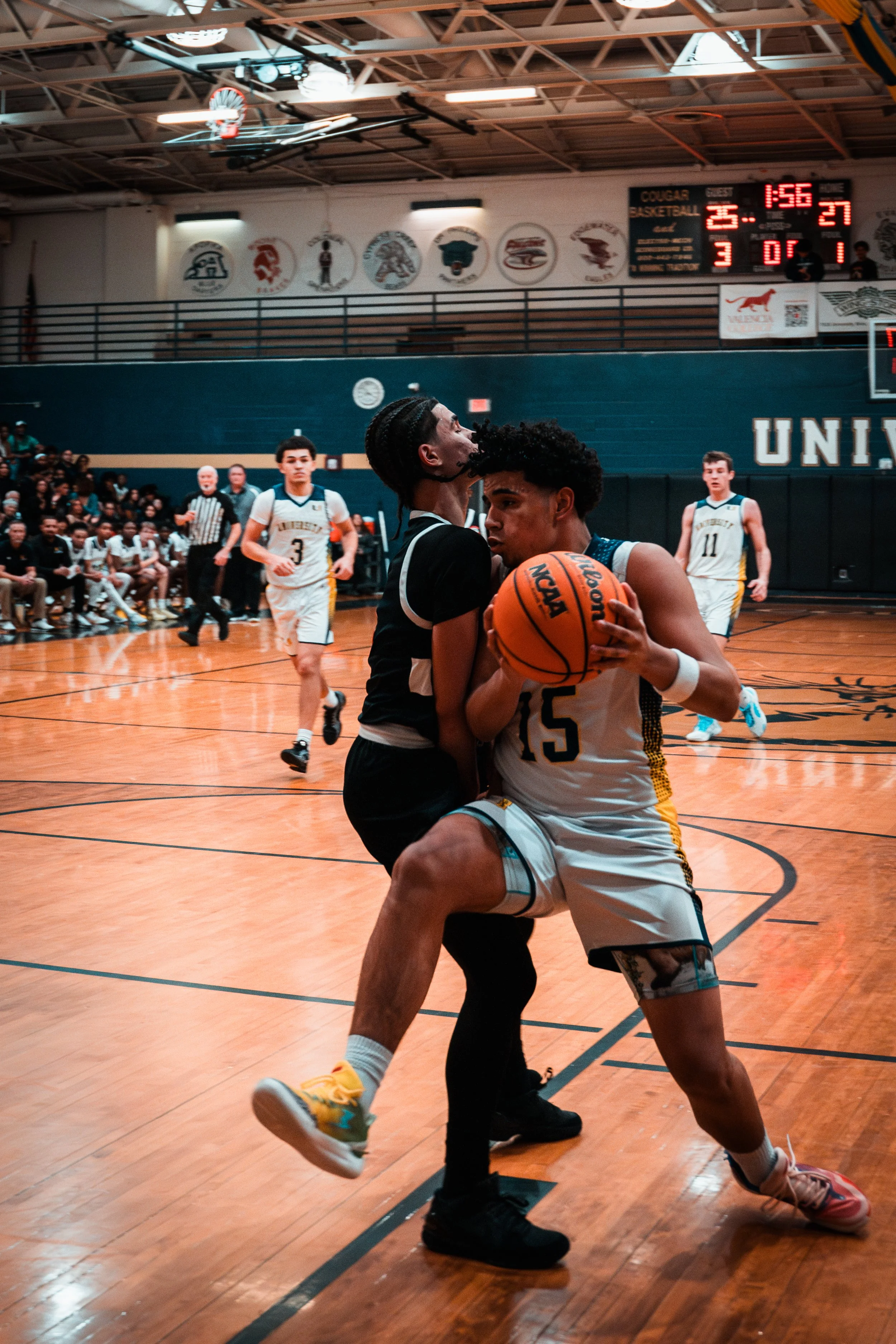 Two basketball players contest for the ball in a game, with one player holding the ball while the other attempts to block. The game is indoors, with a scoreboard showing 1:56 remaining and a score of 25-27. Spectators and other players are visible in