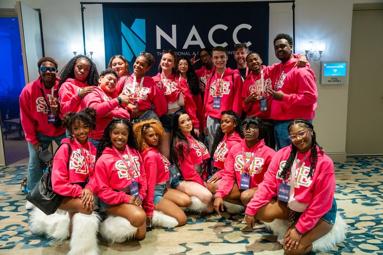 Group of young people and a man wearing pink hoodies with SHE logo, standing in front of a NACCC banner at a conference inside a hotel conference room.