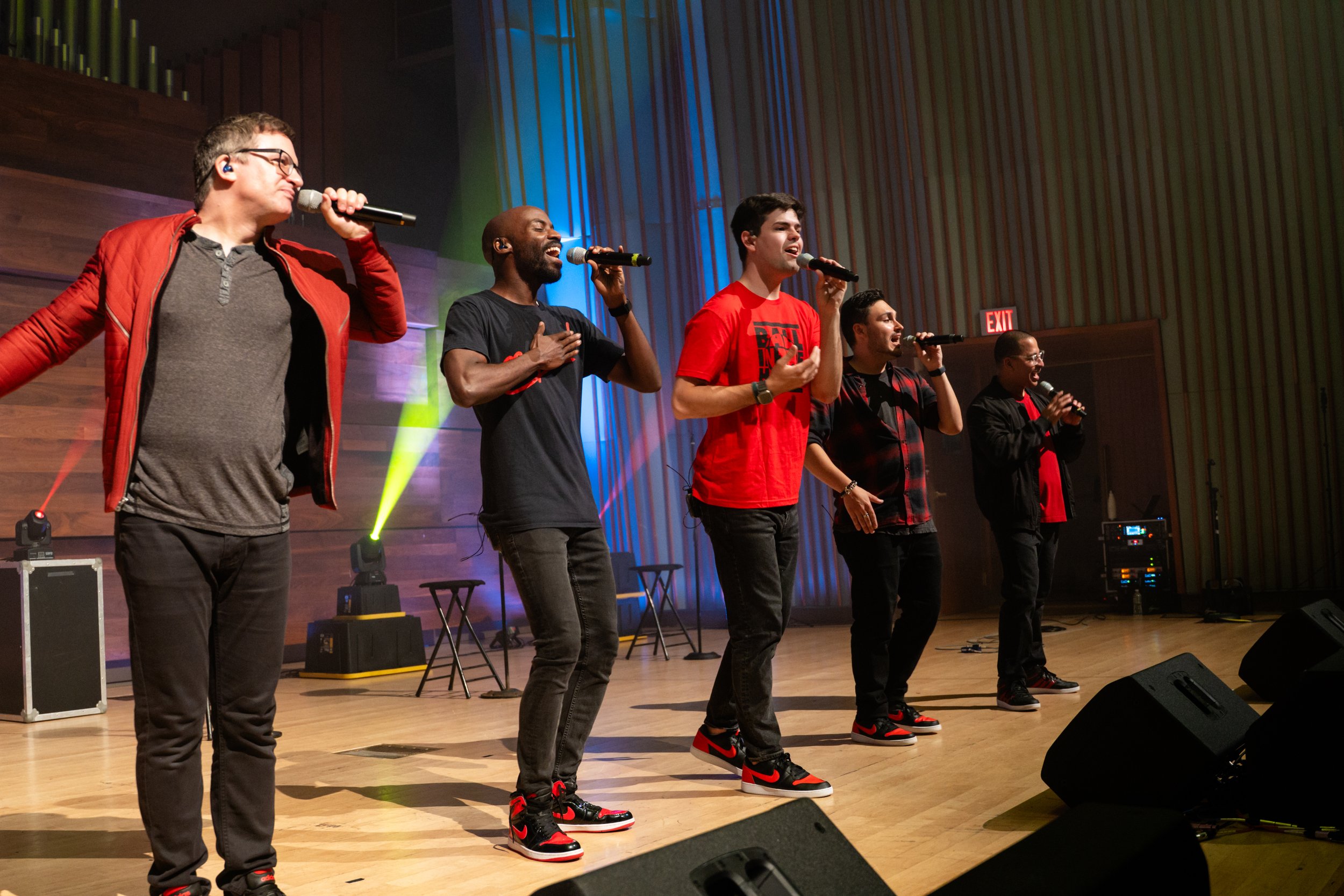 Five men perform on stage with microphones, colorful stage lighting, and sound equipment.