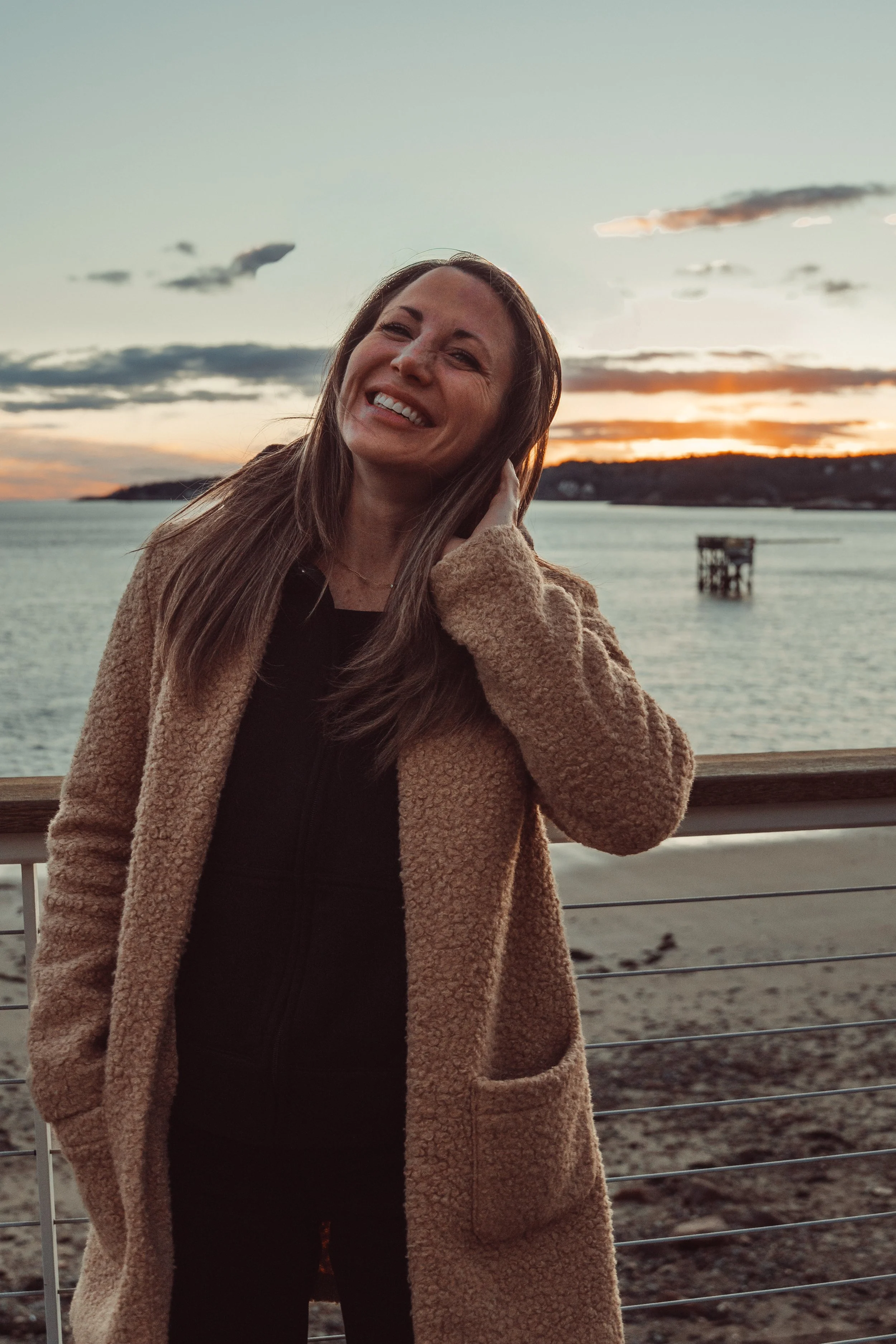 A woman smiling outdoors by a waterfront during sunset, wearing a tan coat and black top.
