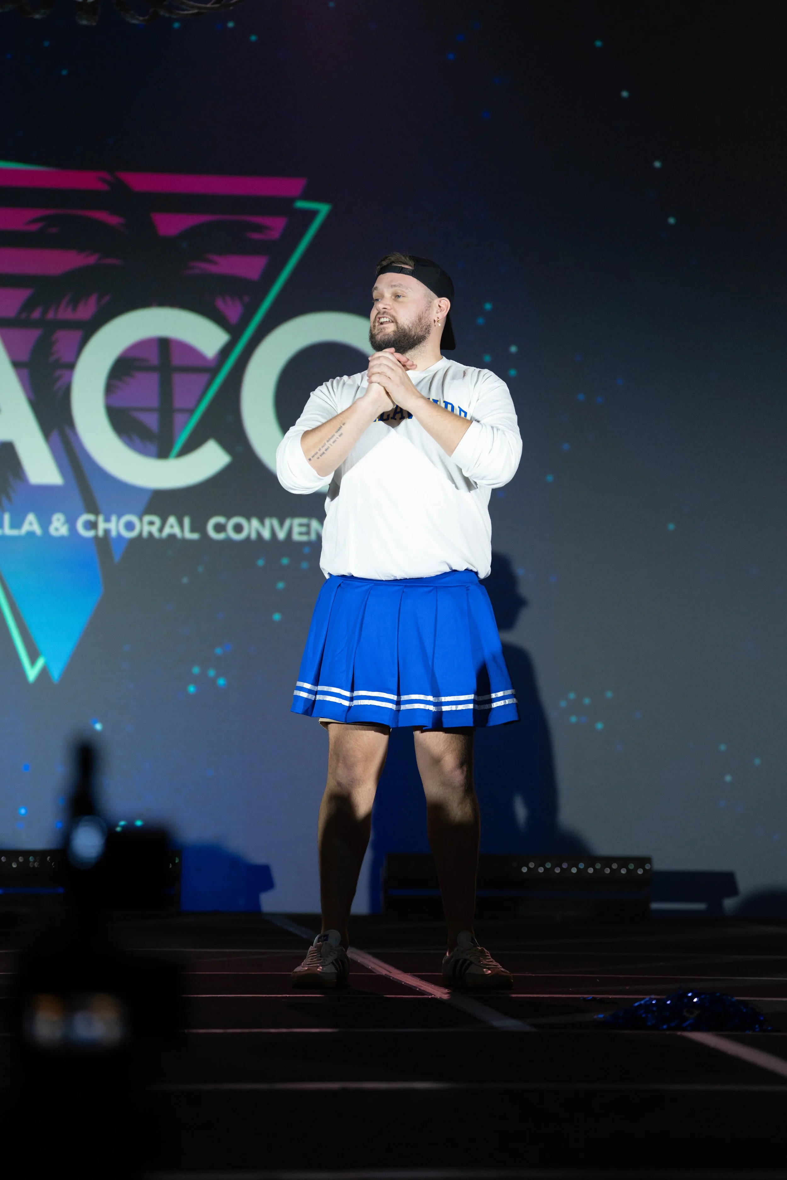 Person with a beard and tattoos on arms, wearing a white shirt, blue skirt, sneakers, and a backwards black cap, standing on a stage in front of a screen with a logo resembling a palm tree and the words 'ACCA' and 'Choral Convention'.