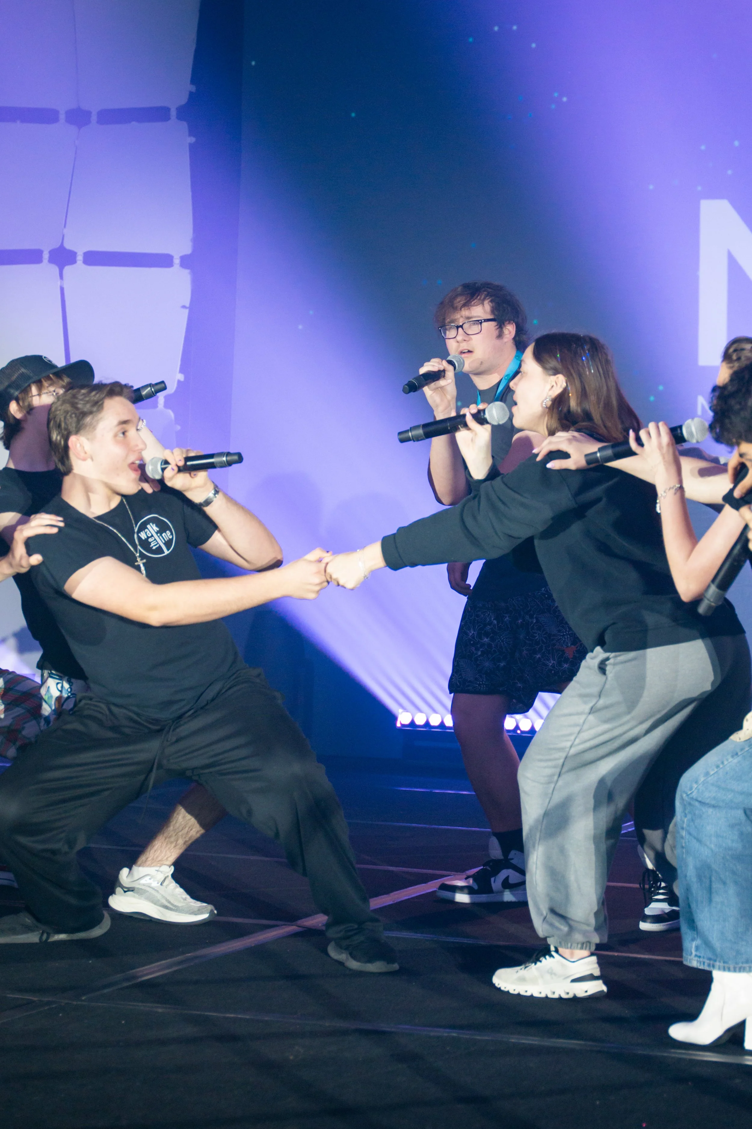 A group of people on stage performing a synchronized dance or routine, holding hands, with a backdrop displaying a digital or cosmic design and stage lighting.