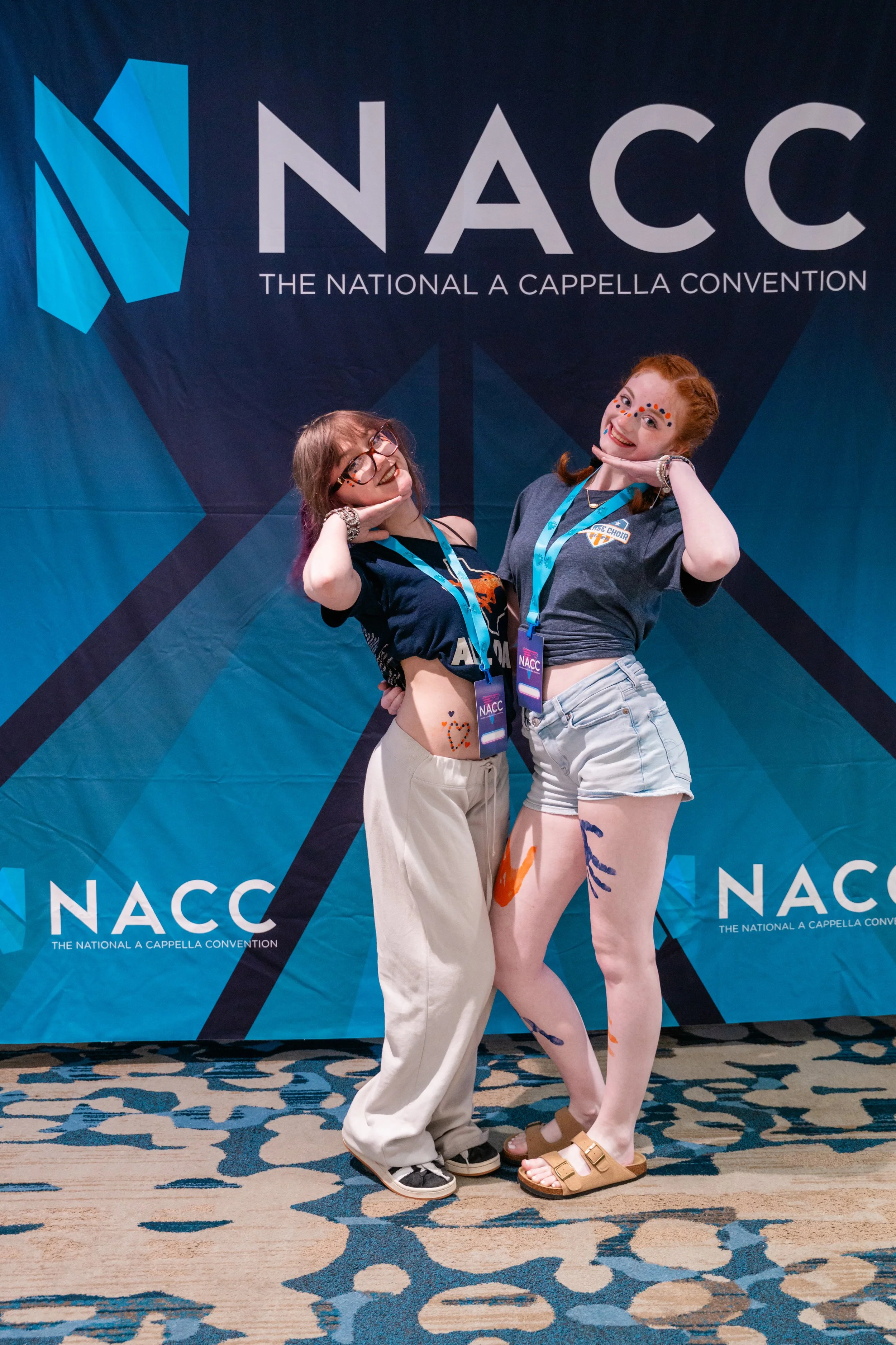 Two women posing in front of a large NACC (The National A Cappella Convention) banner. One woman has glasses and is wearing cream pants and a black shirt, while the other woman has red hair, face paint, and is wearing beige shorts with colorful paint